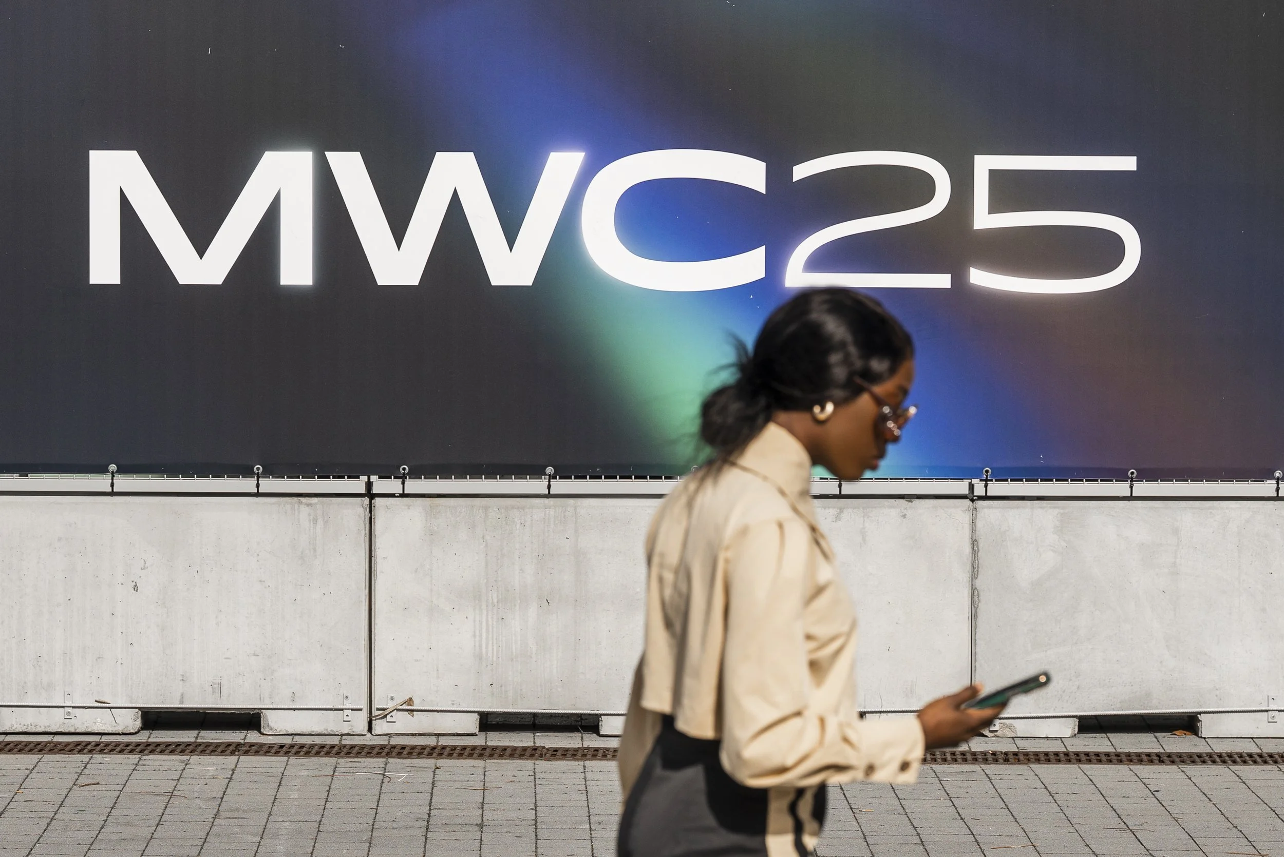A woman walking while looking at her phone in front of a large advertisement board with the text "MWC23".