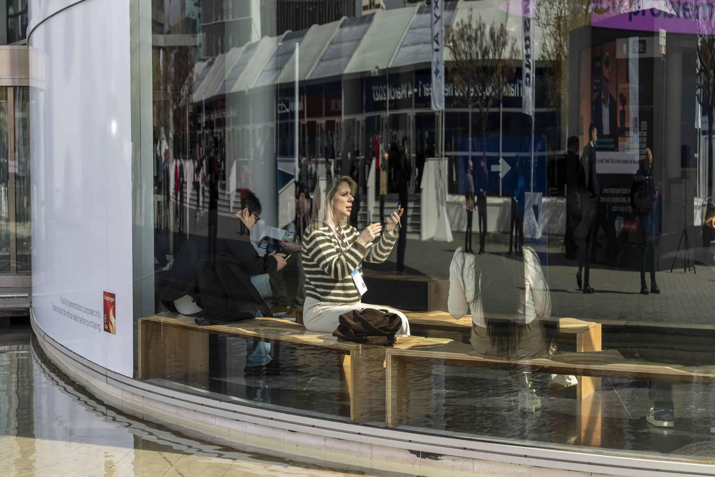 People sitting inside a modern building with large glass windows, with reflections of the outside city scene visible. One woman is engaged with her phone, and another woman is reading a brochure. There are people walking and standing outside visible through the reflections.