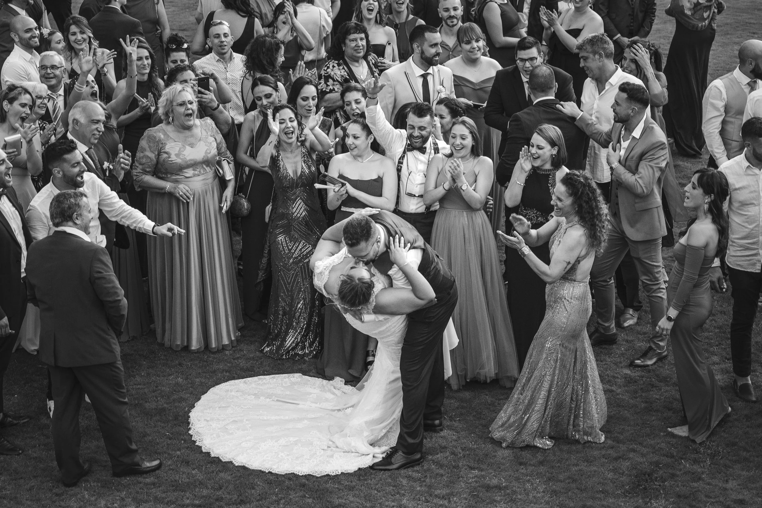 A group of people gathered outdoors at a wedding celebration. The bride and groom are kissing on the dance floor, surrounded by family and friends who are smiling, clapping, and taking photos.