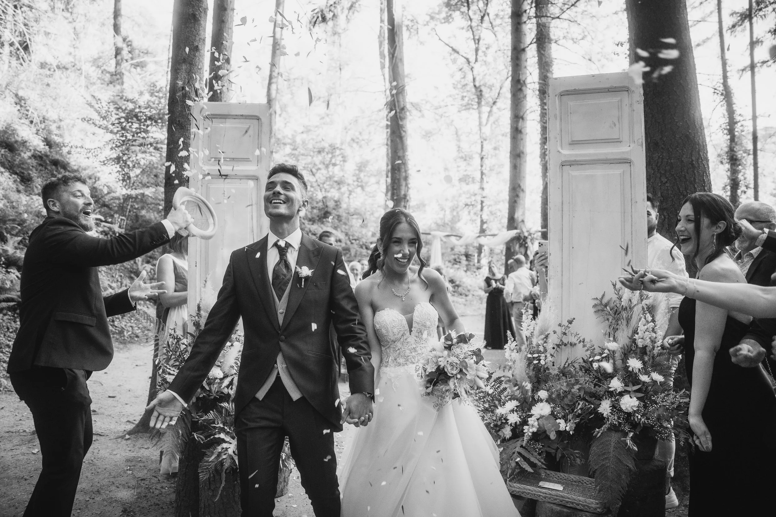 A happy newlywed couple holding hands and smiling, walking through a floral wedding arch in a forest, with friends and family around, celebrating during their outdoor wedding.