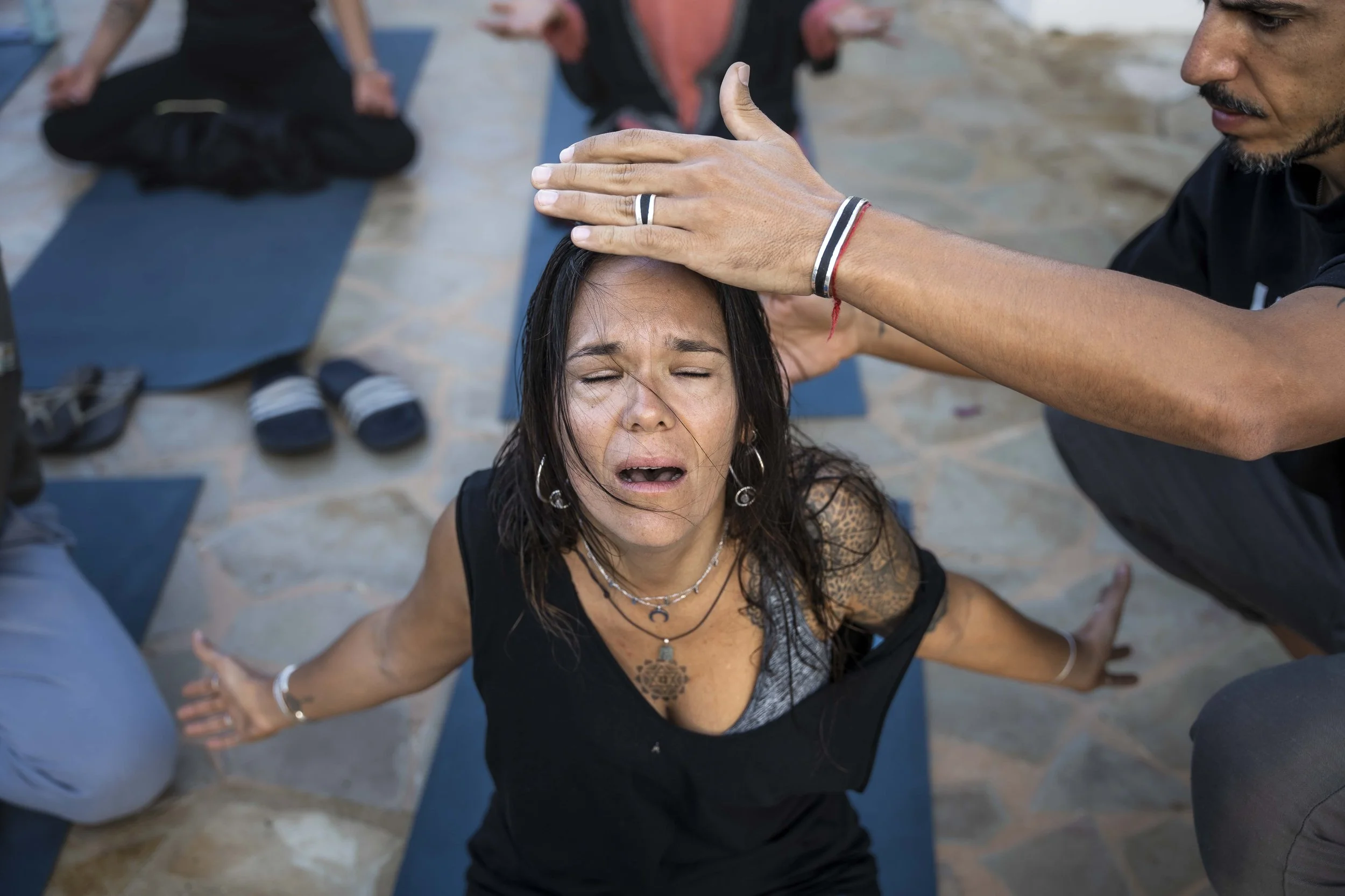 A woman with long dark hair and tattoos on her arms performing a yoga pose outdoors, with a person standing nearby placing a hand on her head in a blessing or healing gesture.