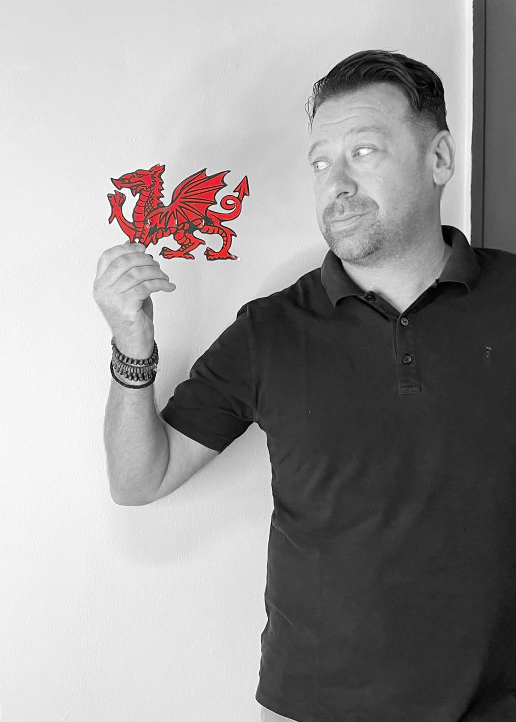 A man with short dark hair and a goatee, wearing a black polo shirt, is holding a red and black dragon sticker in his right hand, looking to the side against a plain white wall.