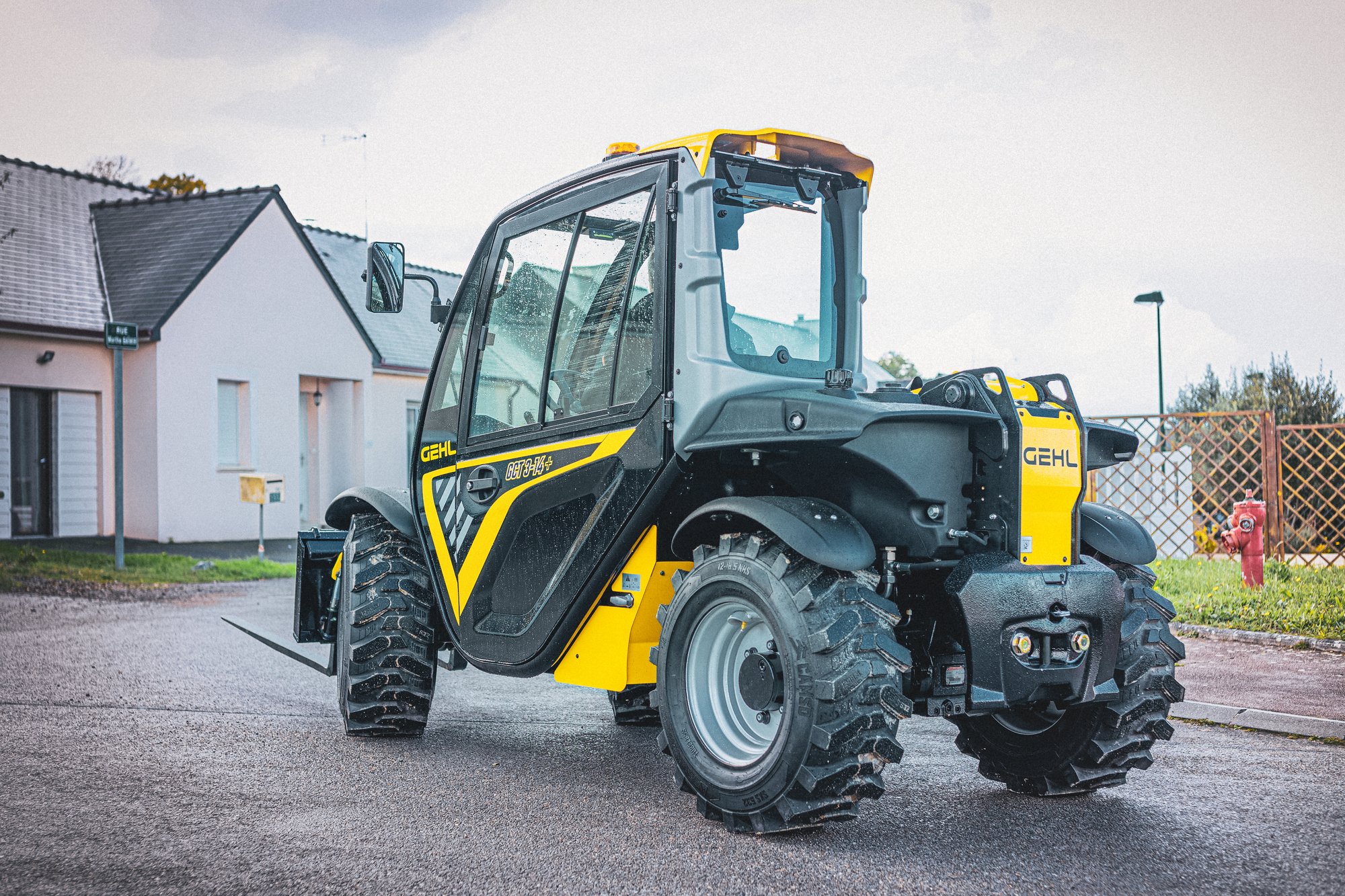 A yellow and black compact construction loader with large rugged tires parked on a street in a residential neighborhood during daylight. Houses with pitched roofs are visible in the background.