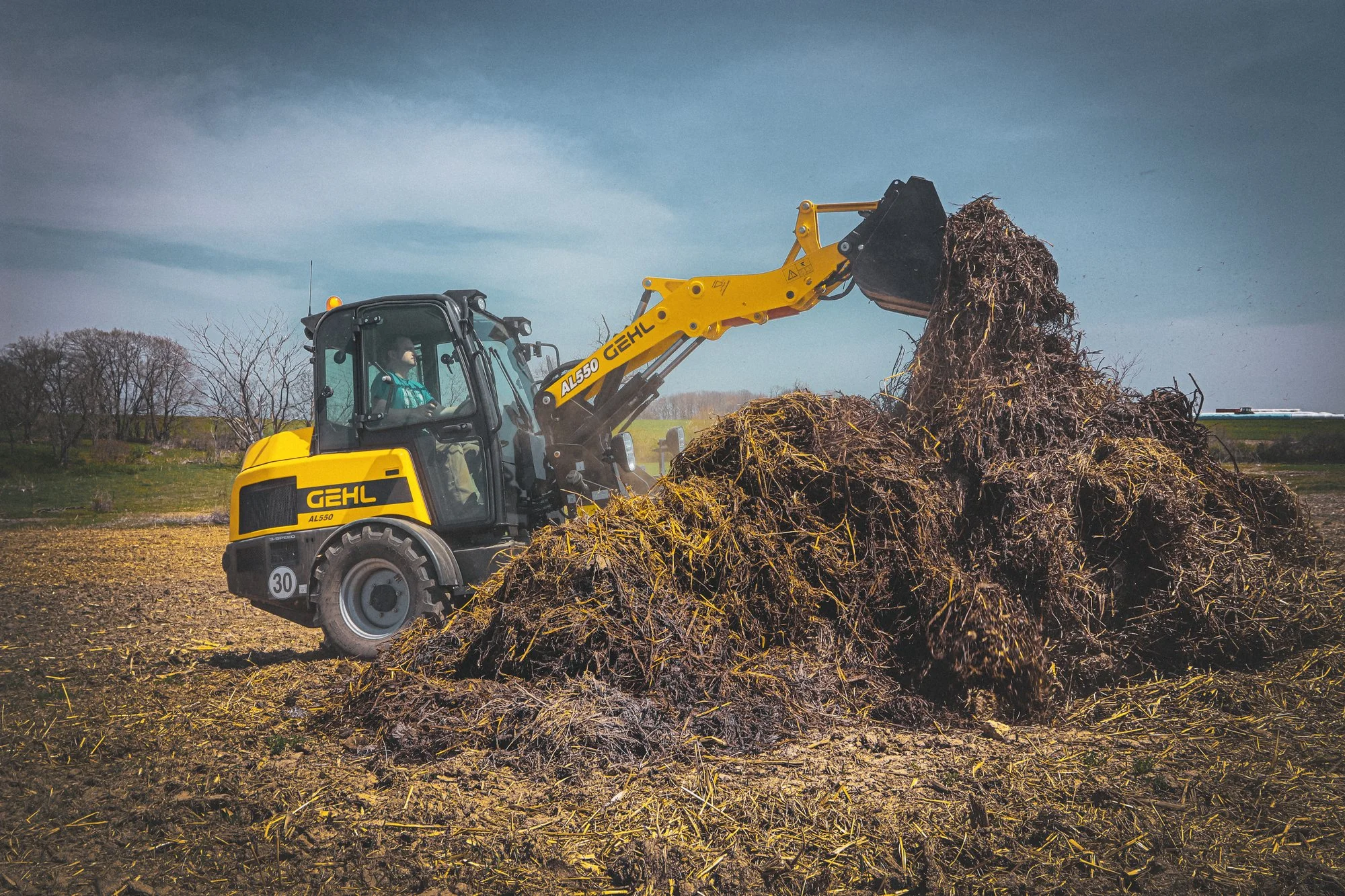 A yellow compact tractor with the brand name 'GEHL' is lifting a large pile of mulch or soil in a field during daytime.