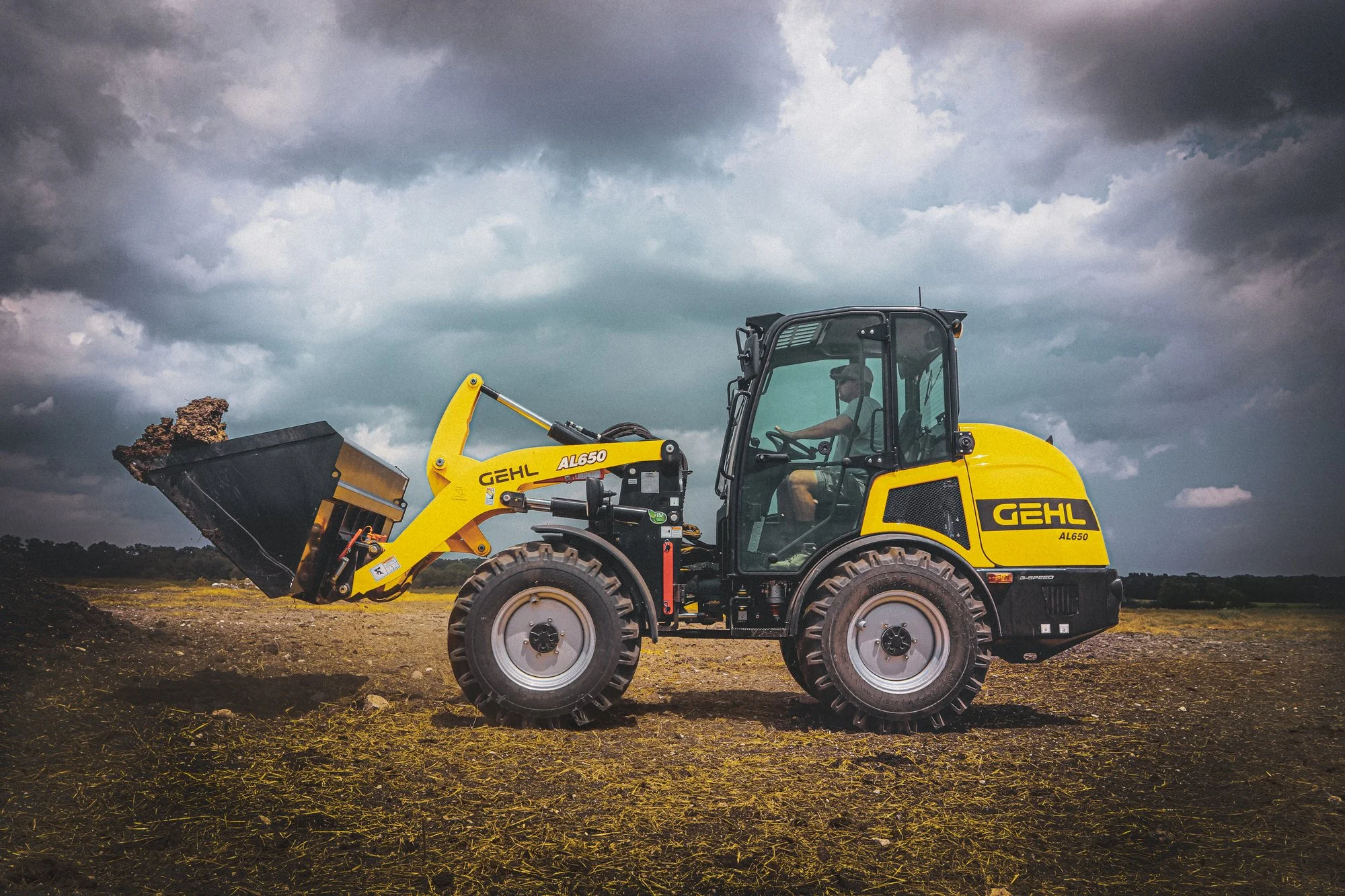 A person operating a yellow Gehl AL650 small loader tractor on a farm, with dark storm clouds overhead and a field of dirt and grass.
