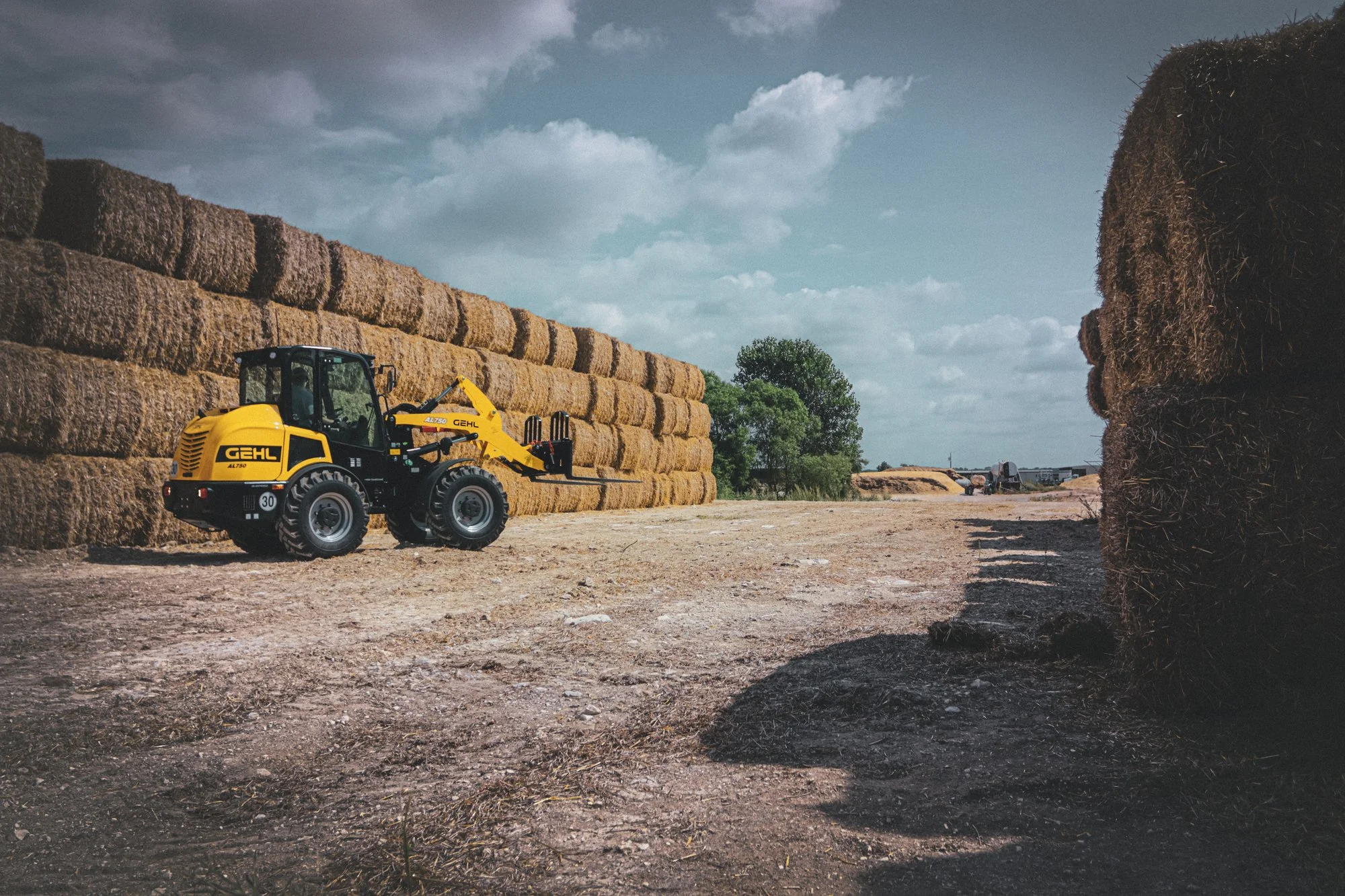 Yellow and black front loader with the brand name GEHL on it, moving hay bales in a farm setting. Large hay bales are stacked on the ground and in the background, with a few trees and a partly cloudy sky.