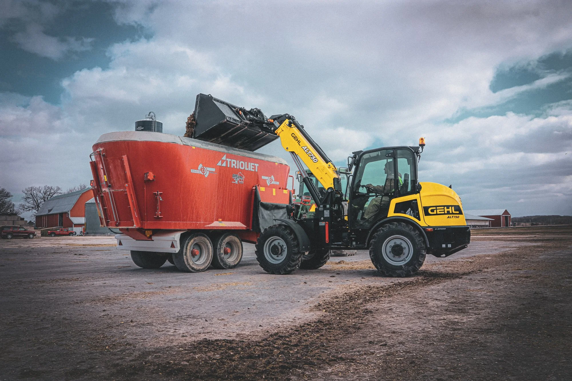 A yellow Gehl AL750 skid steer loader operating on a gravel lot, emptying a black bucket into the red Trioliet feed mixer trailer with a farmhouse and barn in the background.