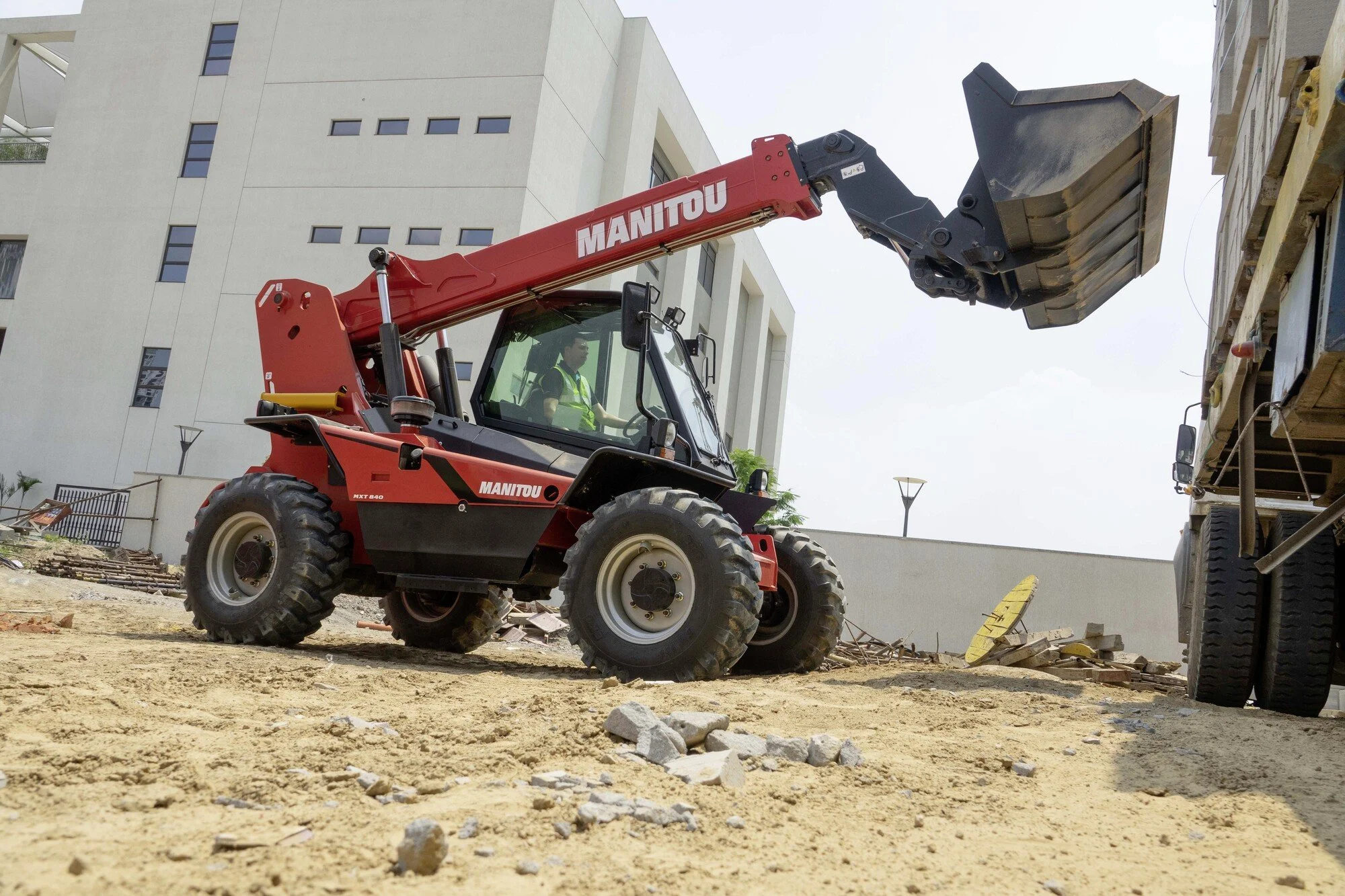 A red Manitou telescopic forklift on a construction site lifting debris. There are modern buildings in the background and a person operating the equipment.