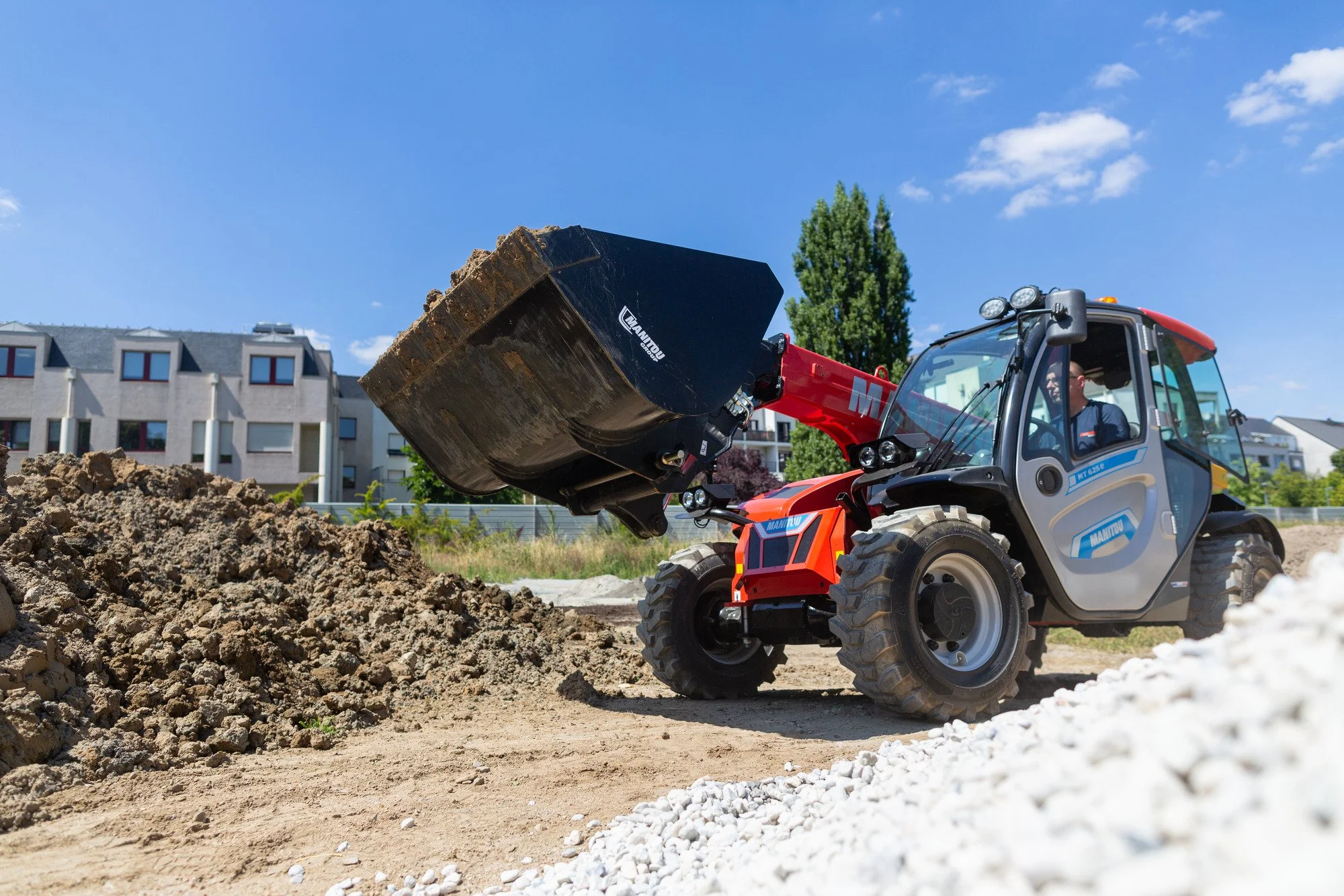 A construction worker operating a small front loader transferring dirt and rocks at a construction site with residential buildings and trees in the background.