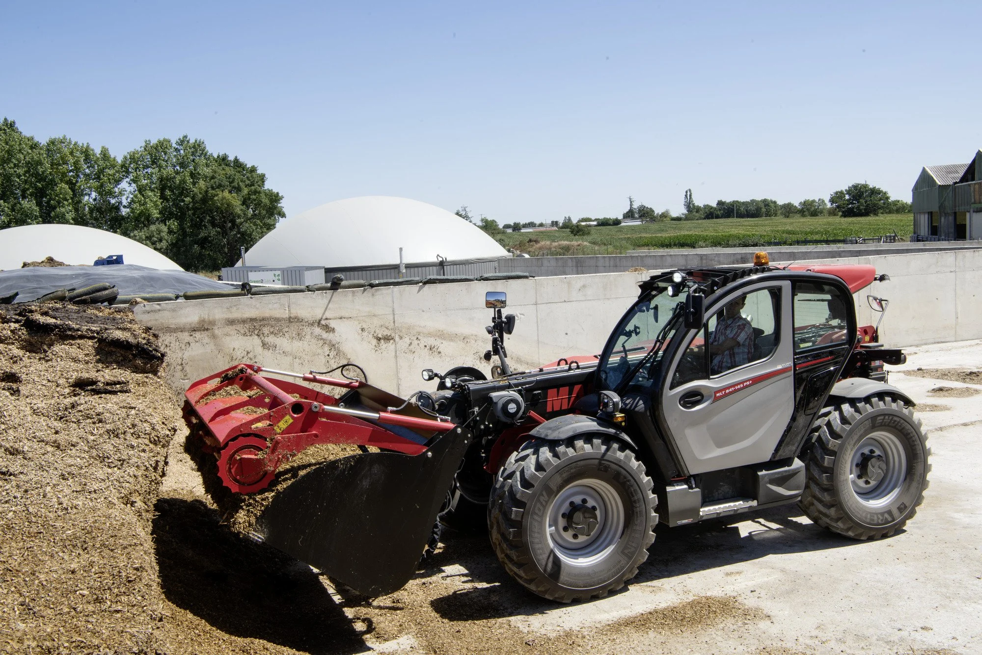 A small tractor with a front loader attachment moving dirt or gravel on a construction site, with large white dome structures and farmland in the background.