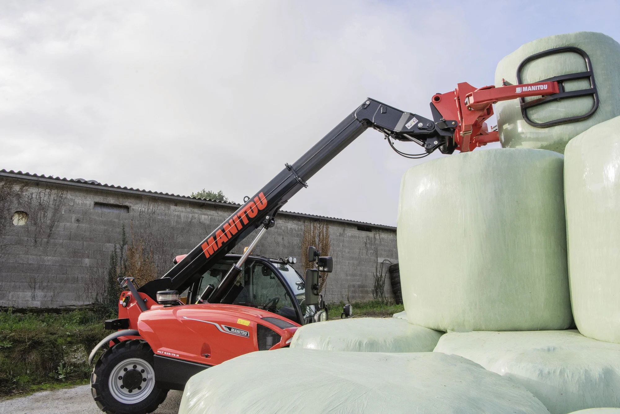 A red Manitou telescopic forklift lifting large white foam blocks outdoors under cloudy sky near a brick wall.