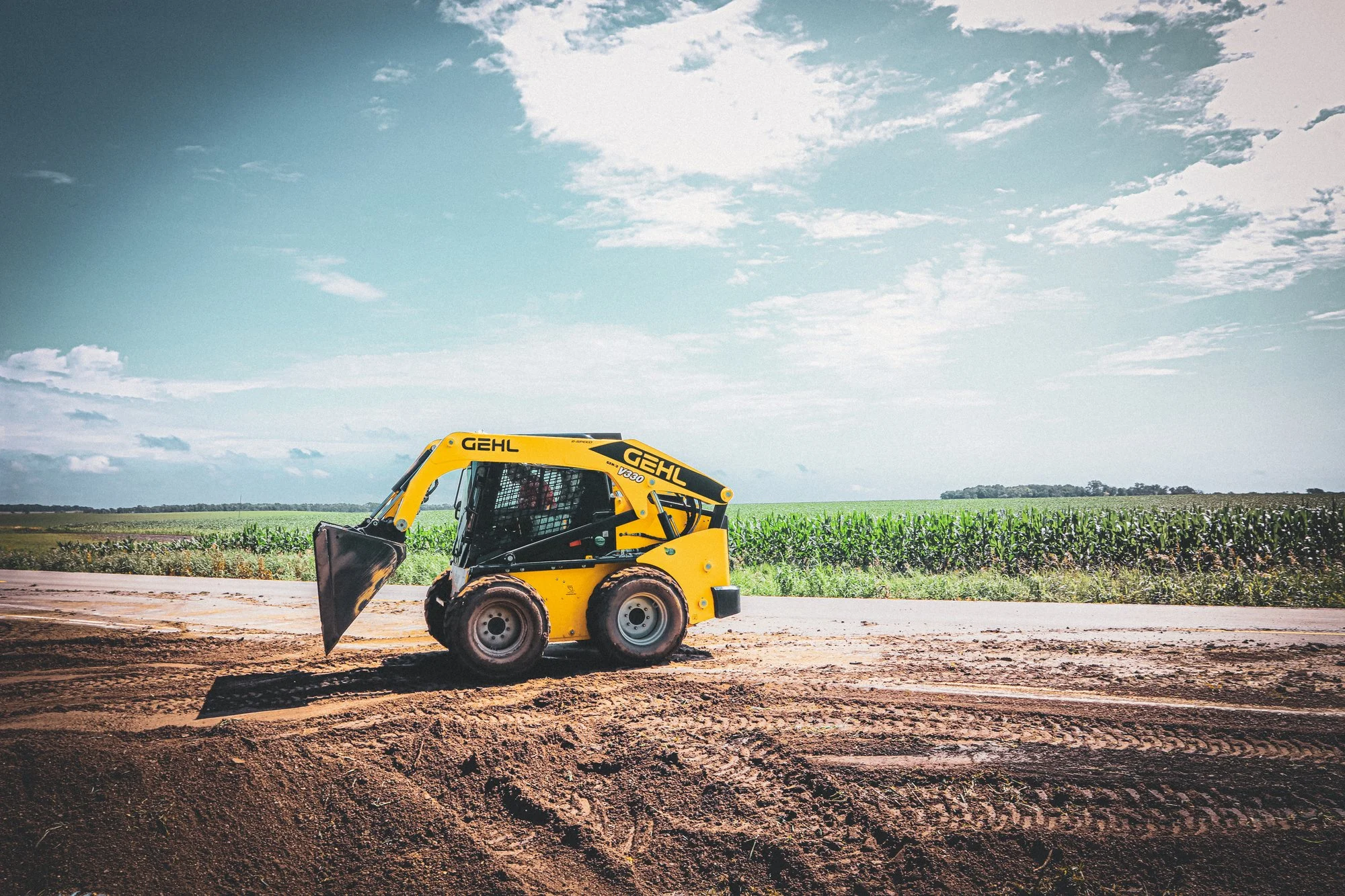 Yellow skid steer loader working on a dirt road beside green crops under partly cloudy blue sky.