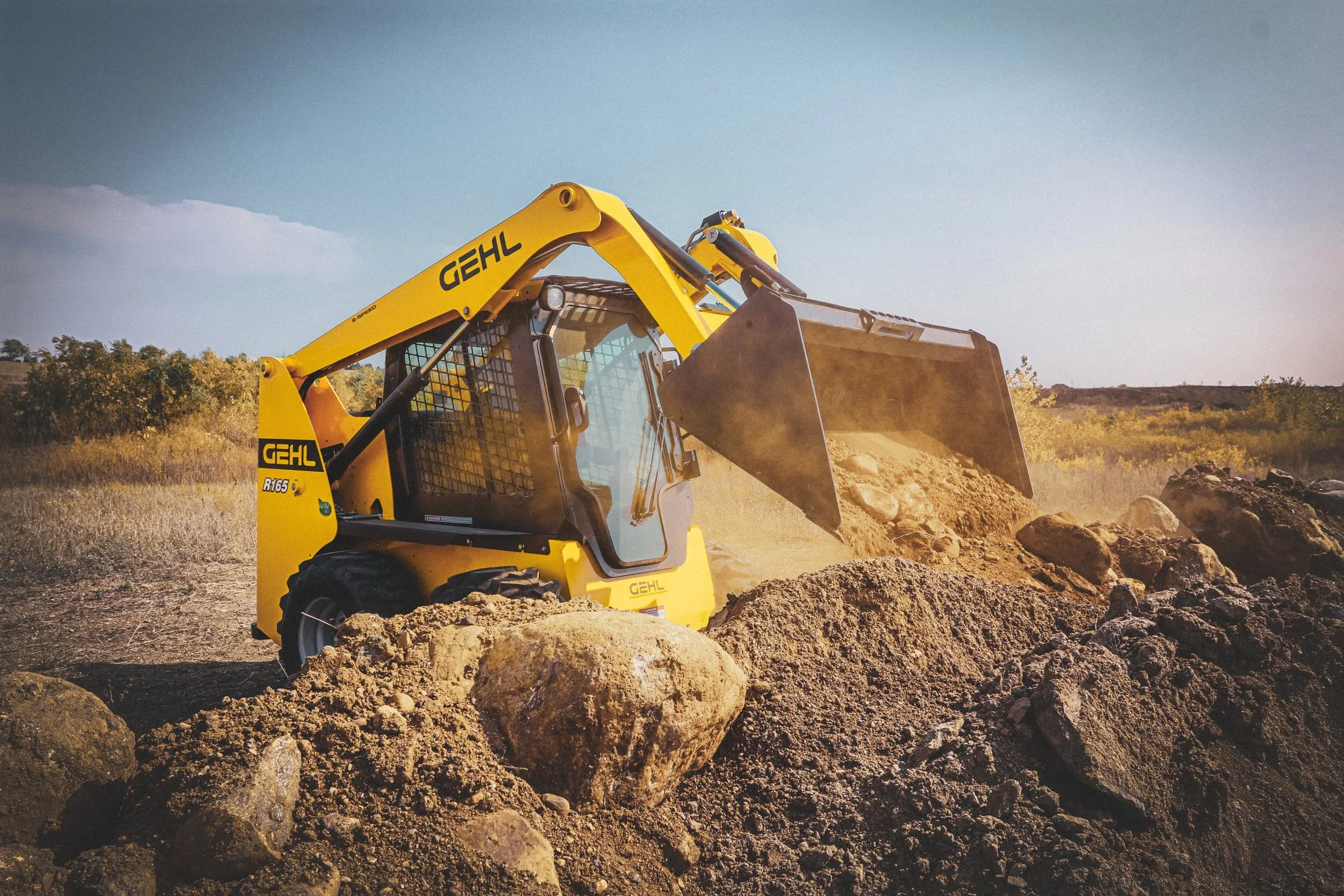 A yellow Gehl compact excavator moving dirt and rocks on an outdoor construction or landscaping site, with trees and a clear blue sky in the background.