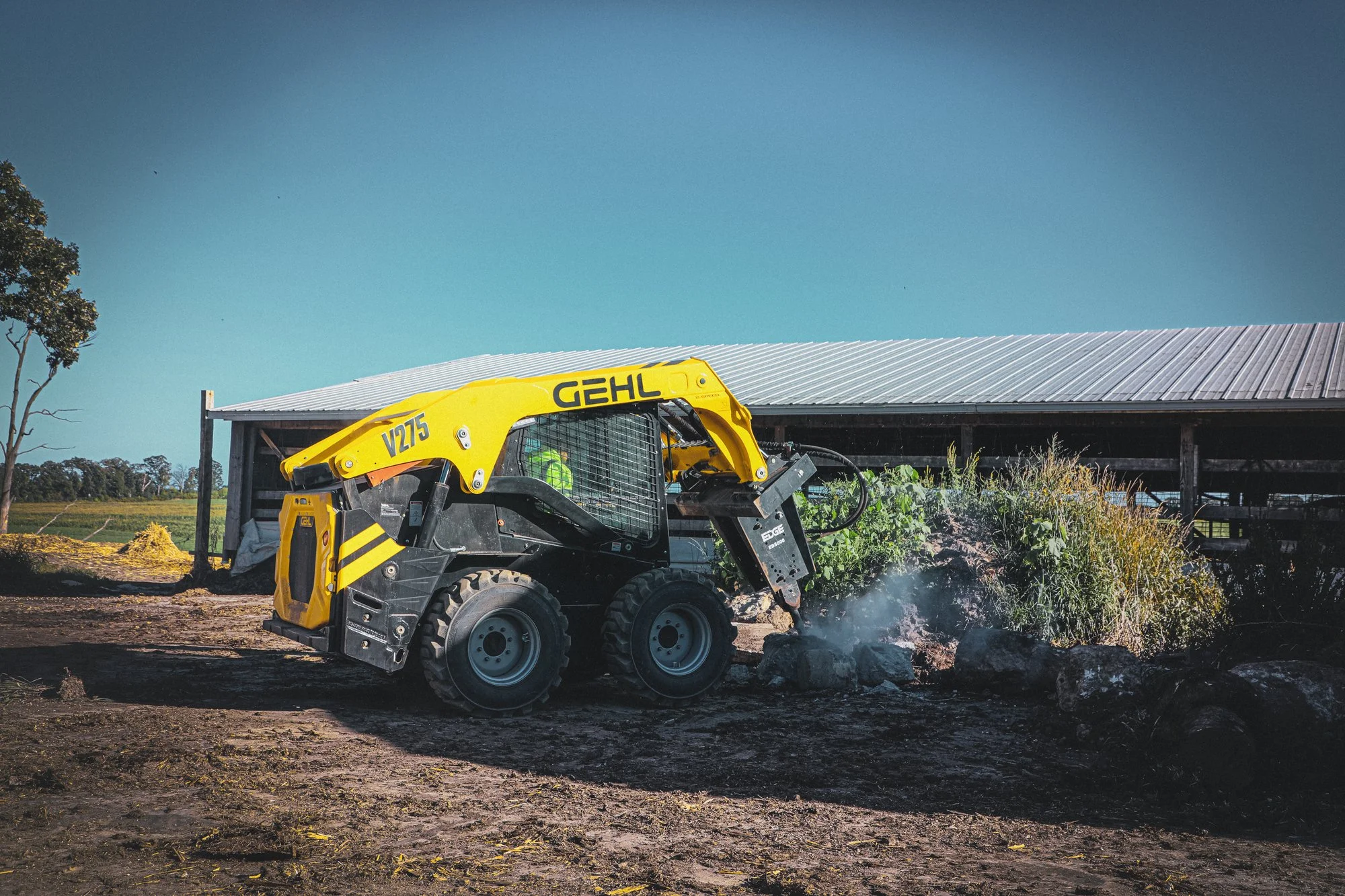 A yellow compact skid steer loader on a construction site, with a building and trees in the background, under a clear blue sky.