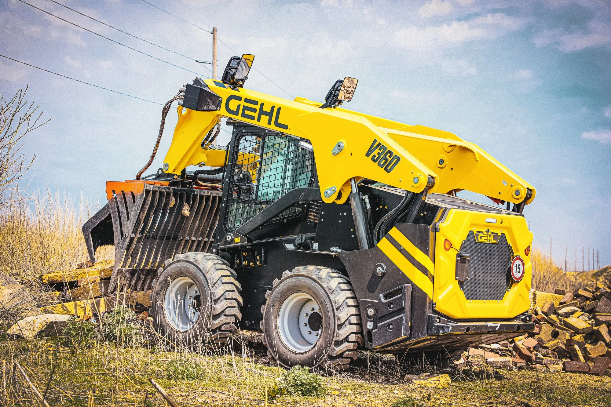 A yellow construction skid steer loader with black accents and the brand name 'GEHL' on the side, operating outdoors on a dirt area with piles of rocks and some sparse grass, under a partly cloudy sky.
