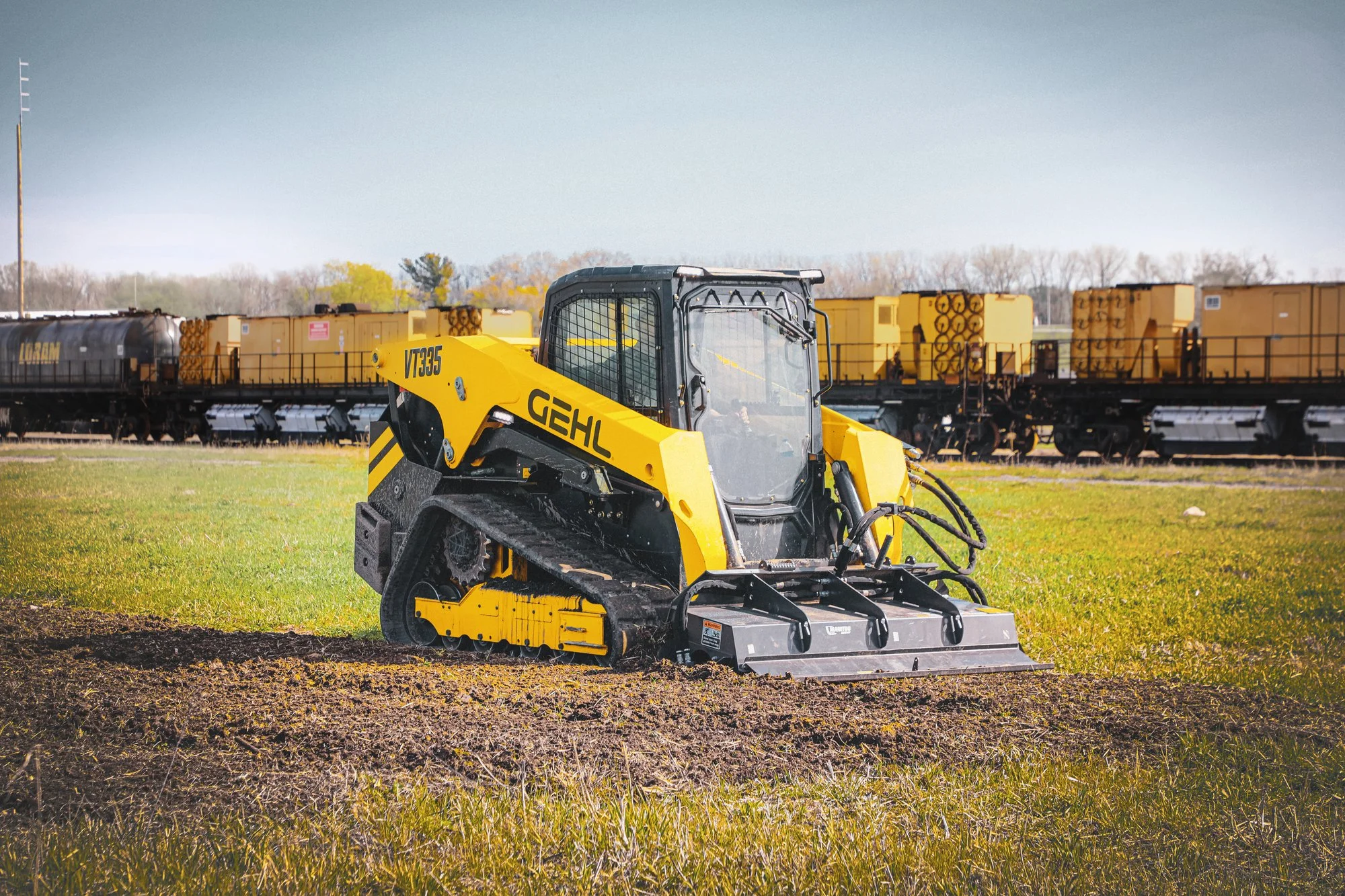 A yellow compact construction vehicle with black tracks and a protective cabin on a grassy field, with train cars on tracks in the background and a blue sky.