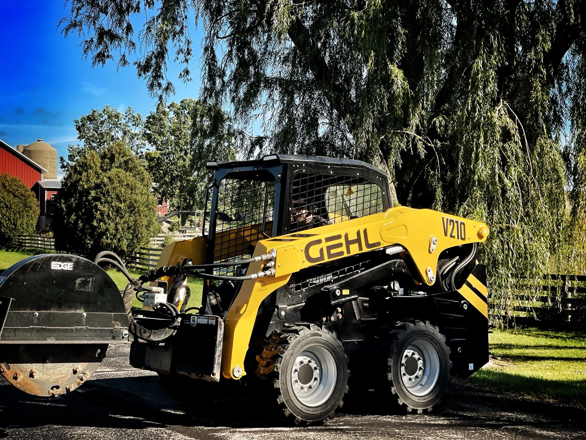 Yellow Gehl V210 skid steer loader parked outdoors with trees, a red barn, and a silo in the background.