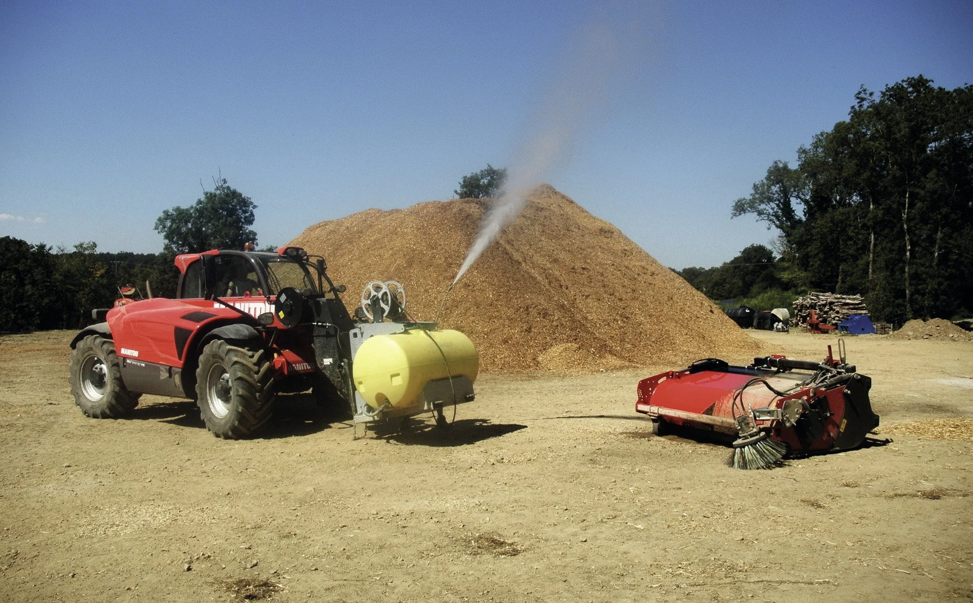 A large pile of wood chips or mulch with a tractor in the foreground, a yellow tank attached to the tractor, and equipment on the ground at an outdoor industrial site under a clear blue sky.