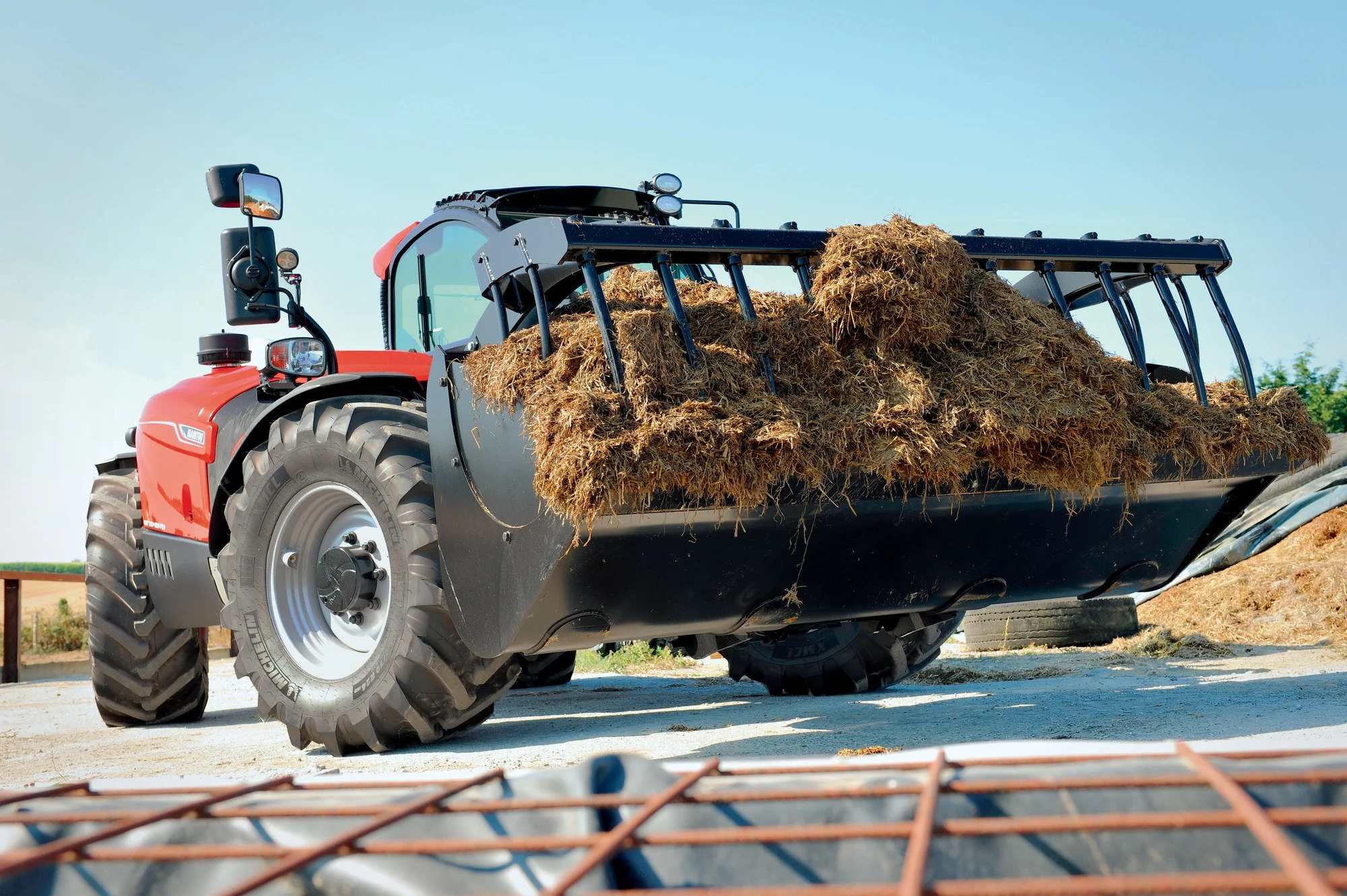 Red tractor with a front loader filled with hay or straw parked outdoors on a farm or field during daytime.