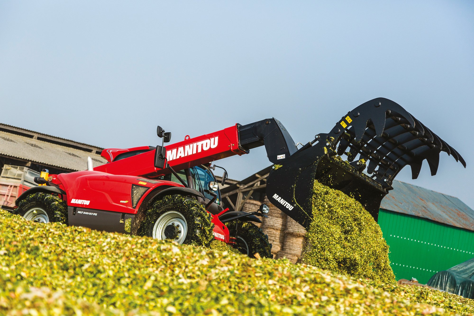 A red Manitou mini loader with a black grapple attachment, working outdoors on a farm or rural property. The background shows farm buildings and a clear blue sky.