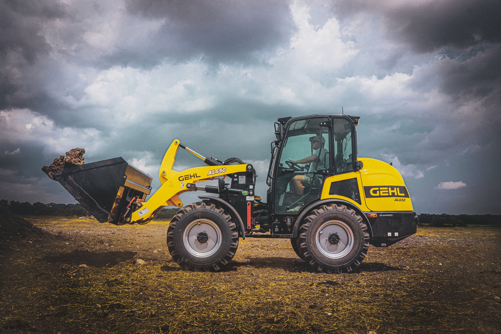 A yellow Gehl AL650 compact loader operating on a farm field during cloudy weather.
