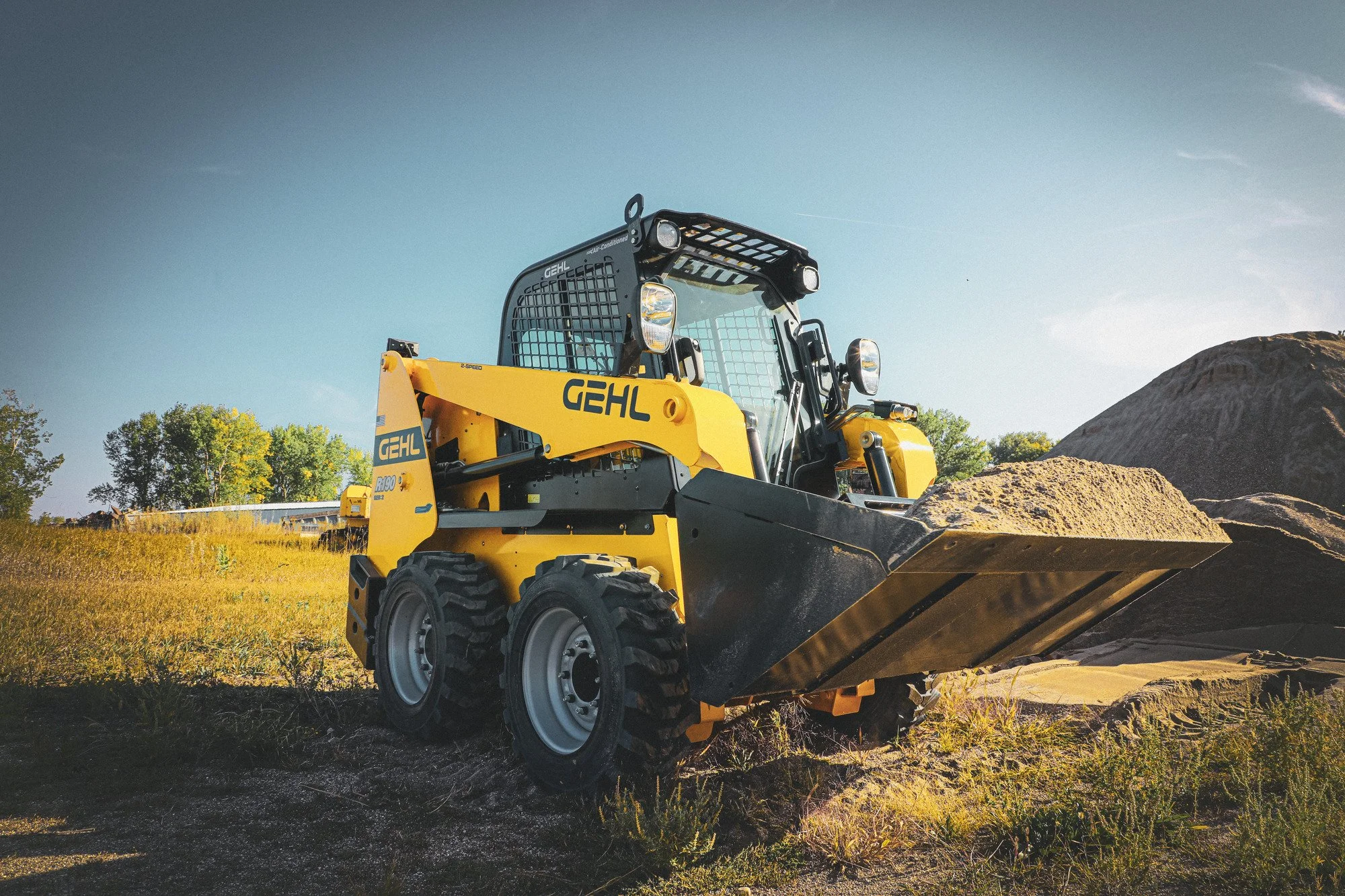 Yellow gehl compact excavator with a large bucket attached, situated outdoors on a construction site with a mound of dirt, grass, and trees in the background, under a clear blue sky.