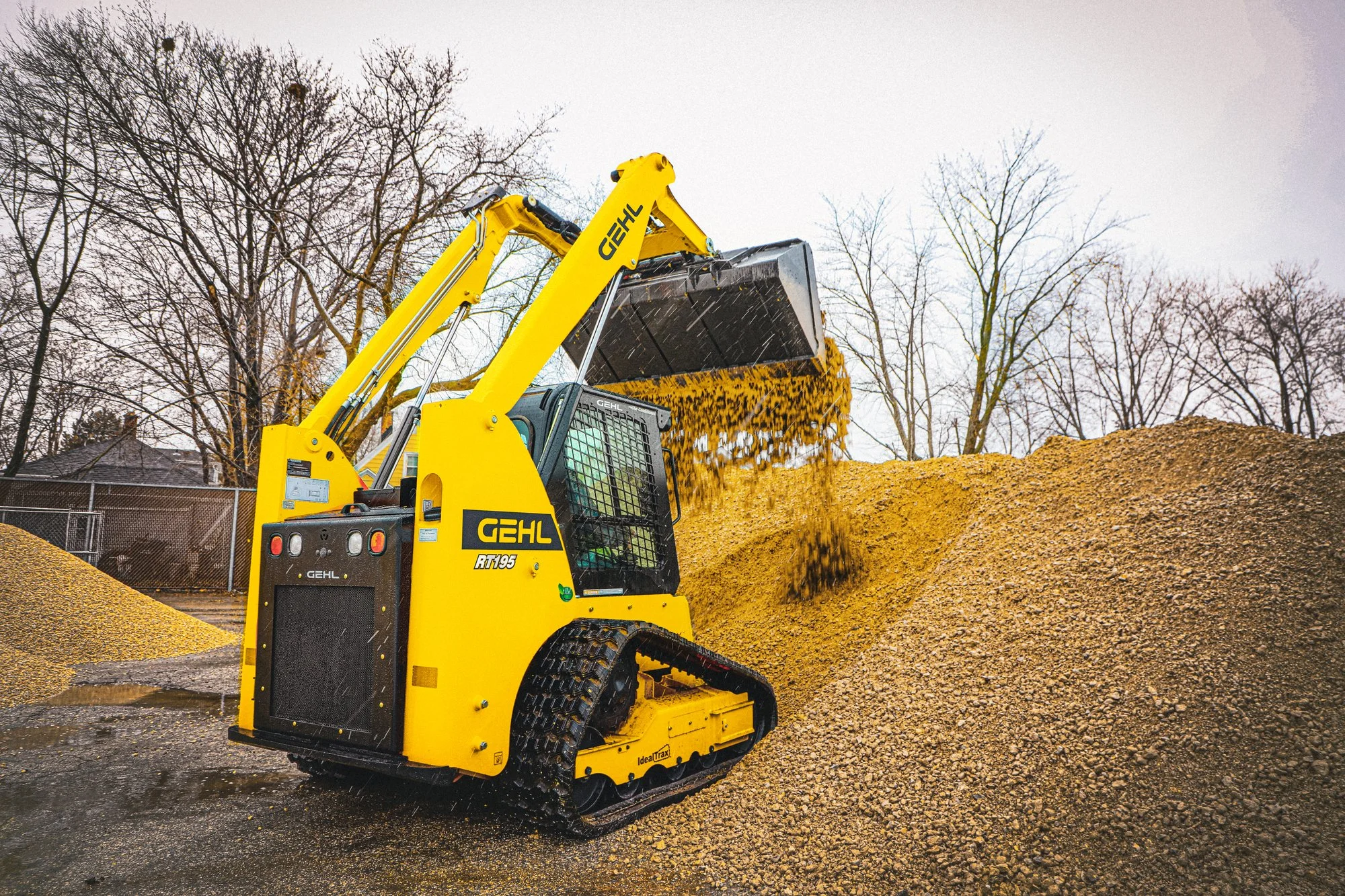 A yellow Gehl RT195 compact excavator with rubber tracks loading a large pile of gravel or soil outdoors during winter or early spring, with bare trees and overcast sky in the background.