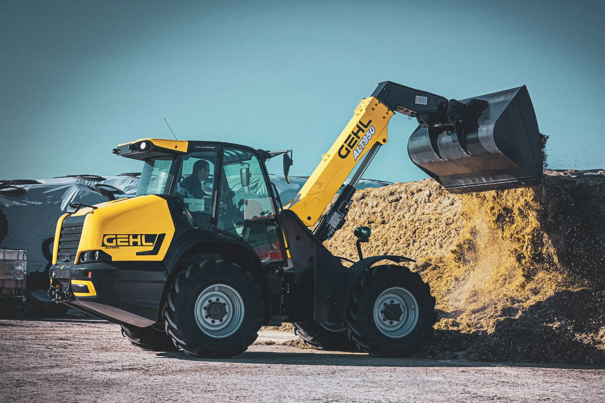 A yellow Gehl compact wheel loader moving a pile of dirt or mulch at a construction or landscaping site.