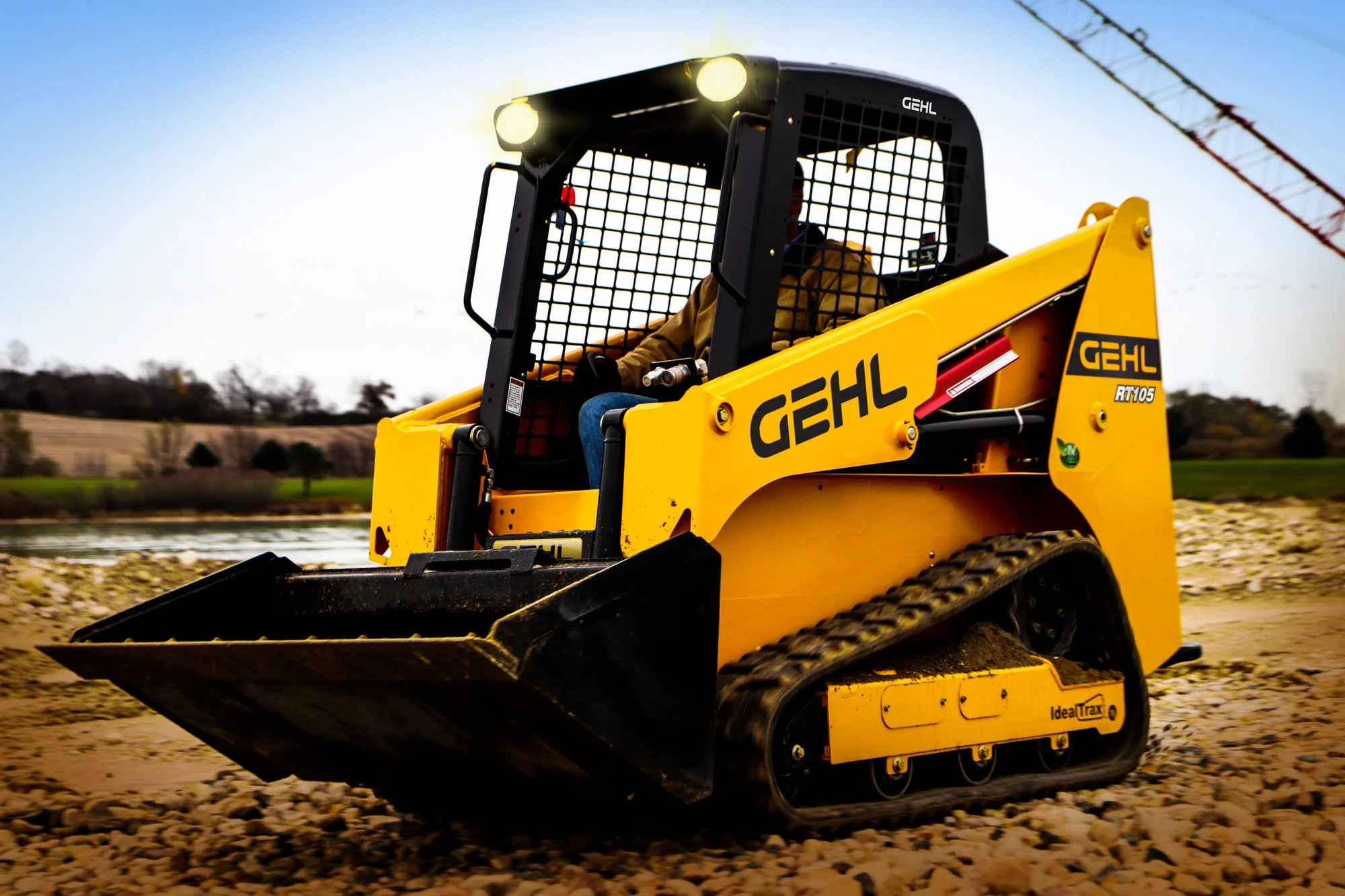 Yellow GEHL skid-steer loader working on a dirt and gravel site with a crane in the background under a clear blue sky.