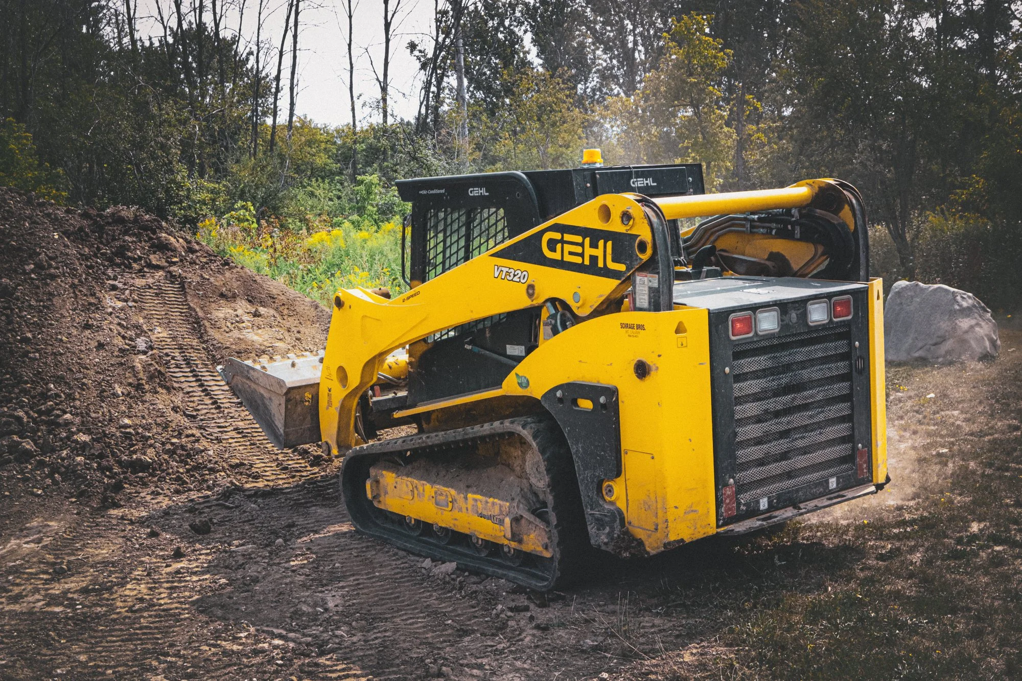Yellow and black compact excavator on dirt construction site with trees in background.