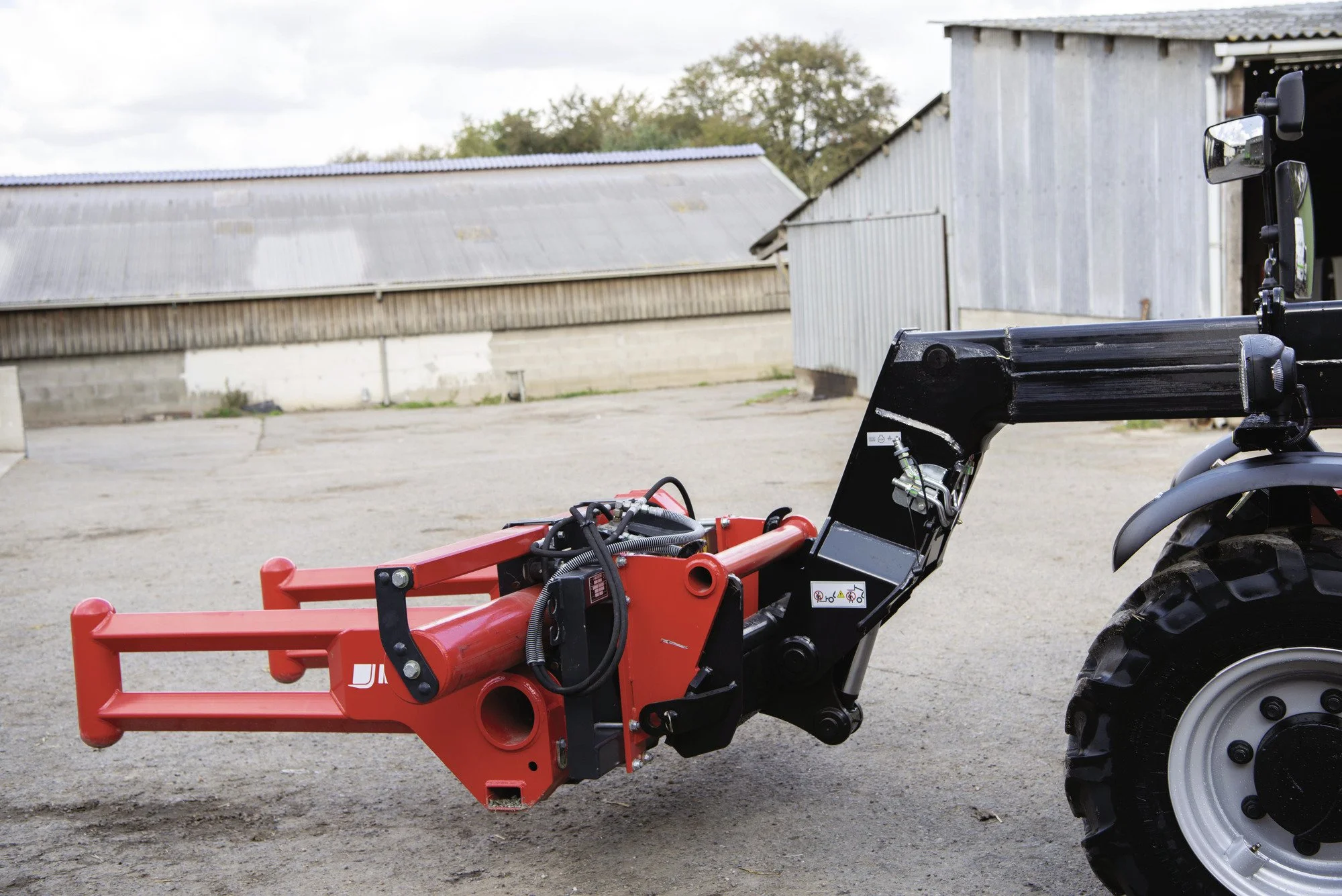 A tractor with a red implement attached to the front, parked on a farmyard with old metal sheds in the background under a cloudy sky.