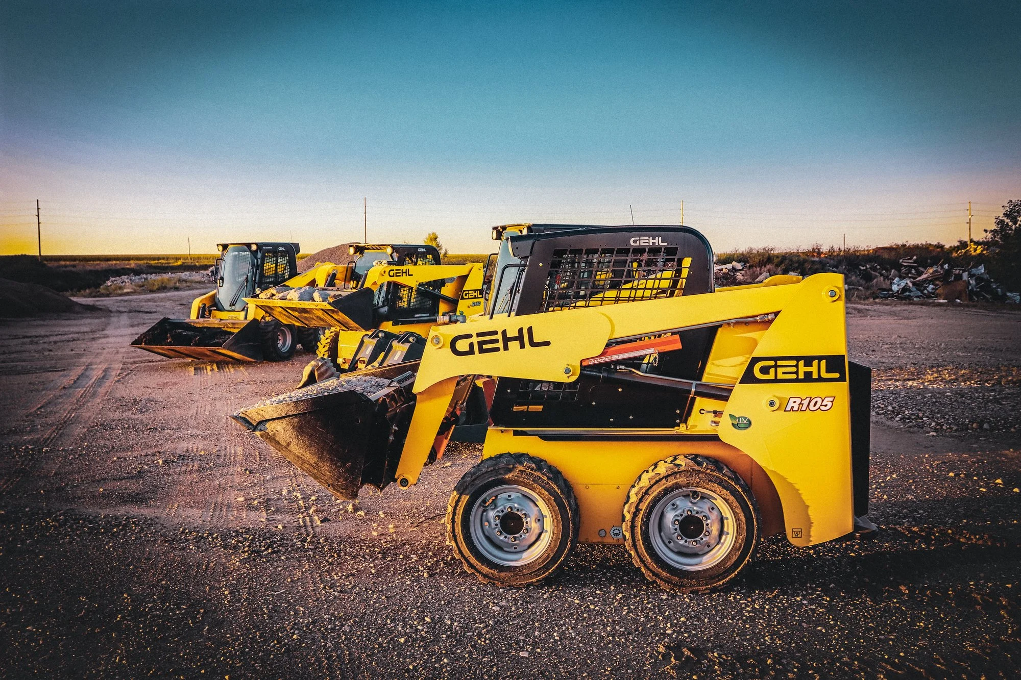 A row of yellow gehl skid-steer loaders parked on a dirt surface at sunset