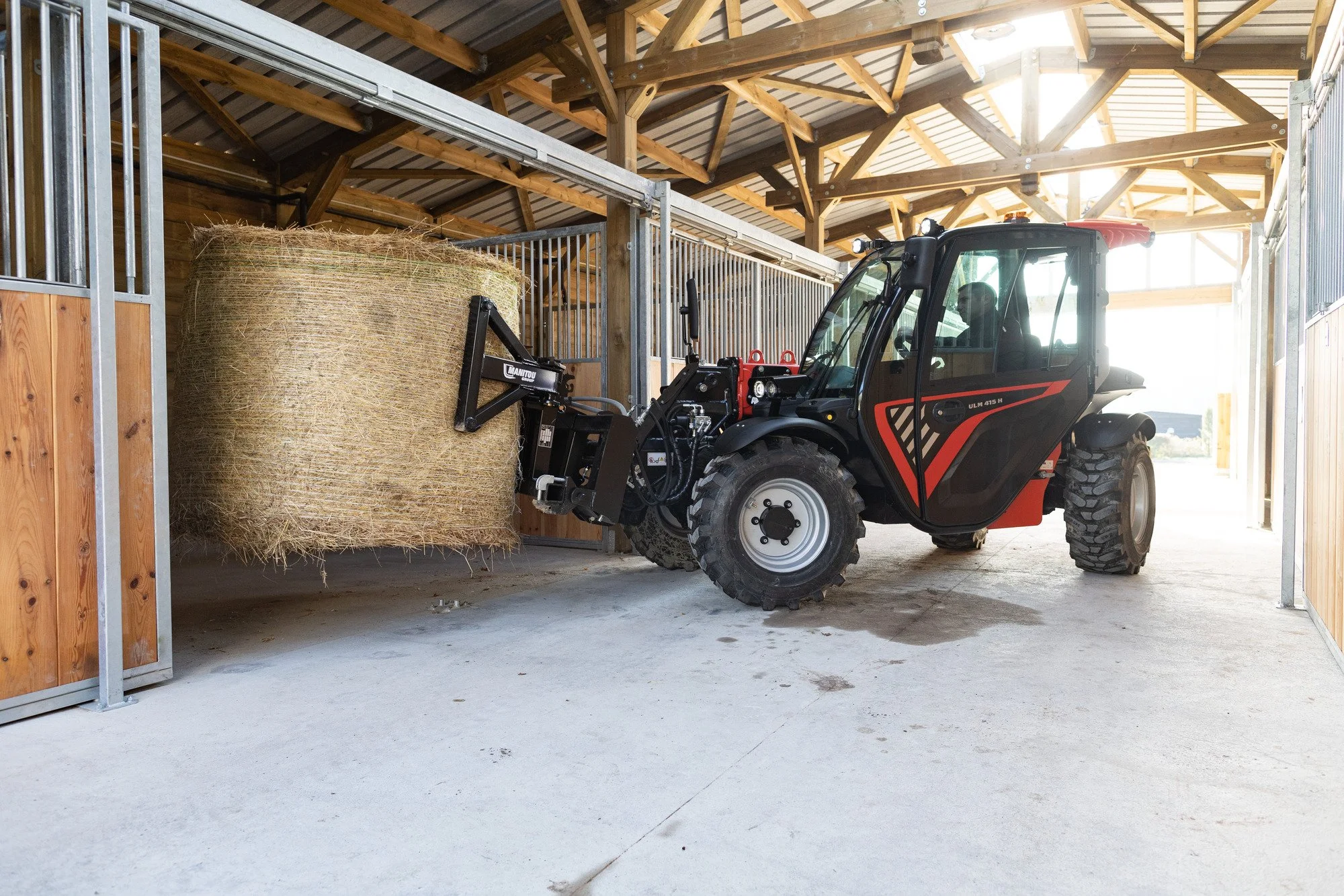 A red and black utility vehicle lifting a large hay bale inside a wooden barn with a metal roof.