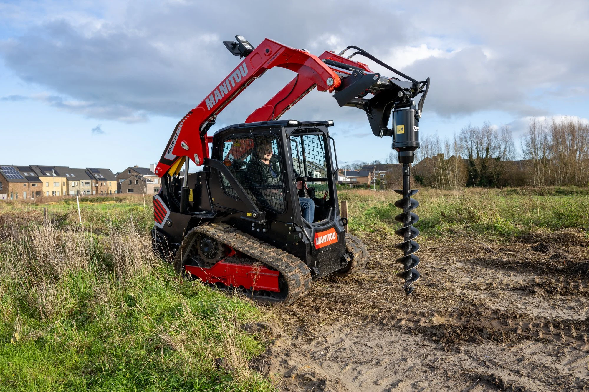 A person operating a red Manitou compact mini excavator with a black hydraulic drill attachment on a construction site with houses and trees in the background under partly cloudy sky.