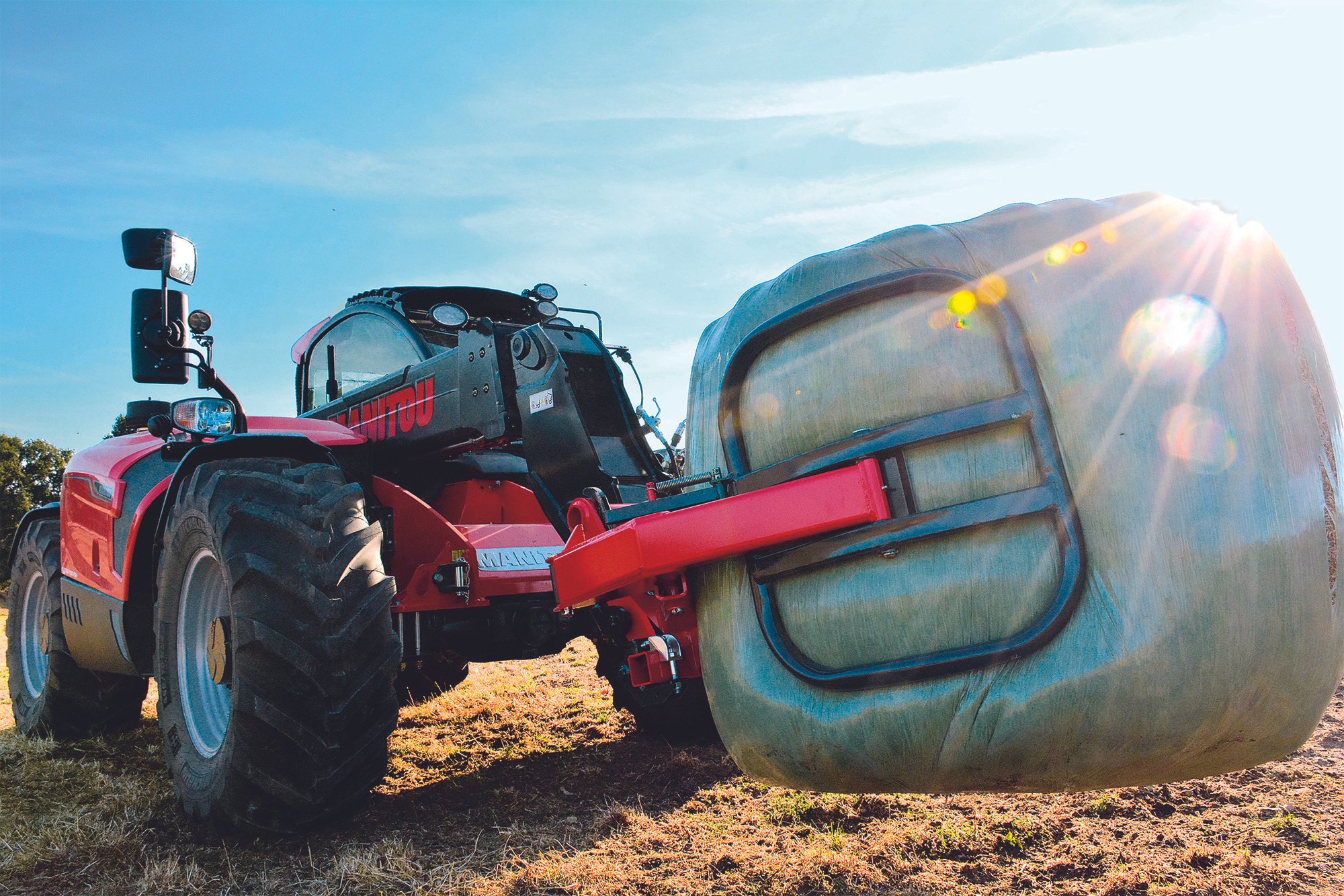 A red tractor with a large tank attached, likely for liquid spreading, situated on a grassy field under a blue sky with sunlight flaring to the right.