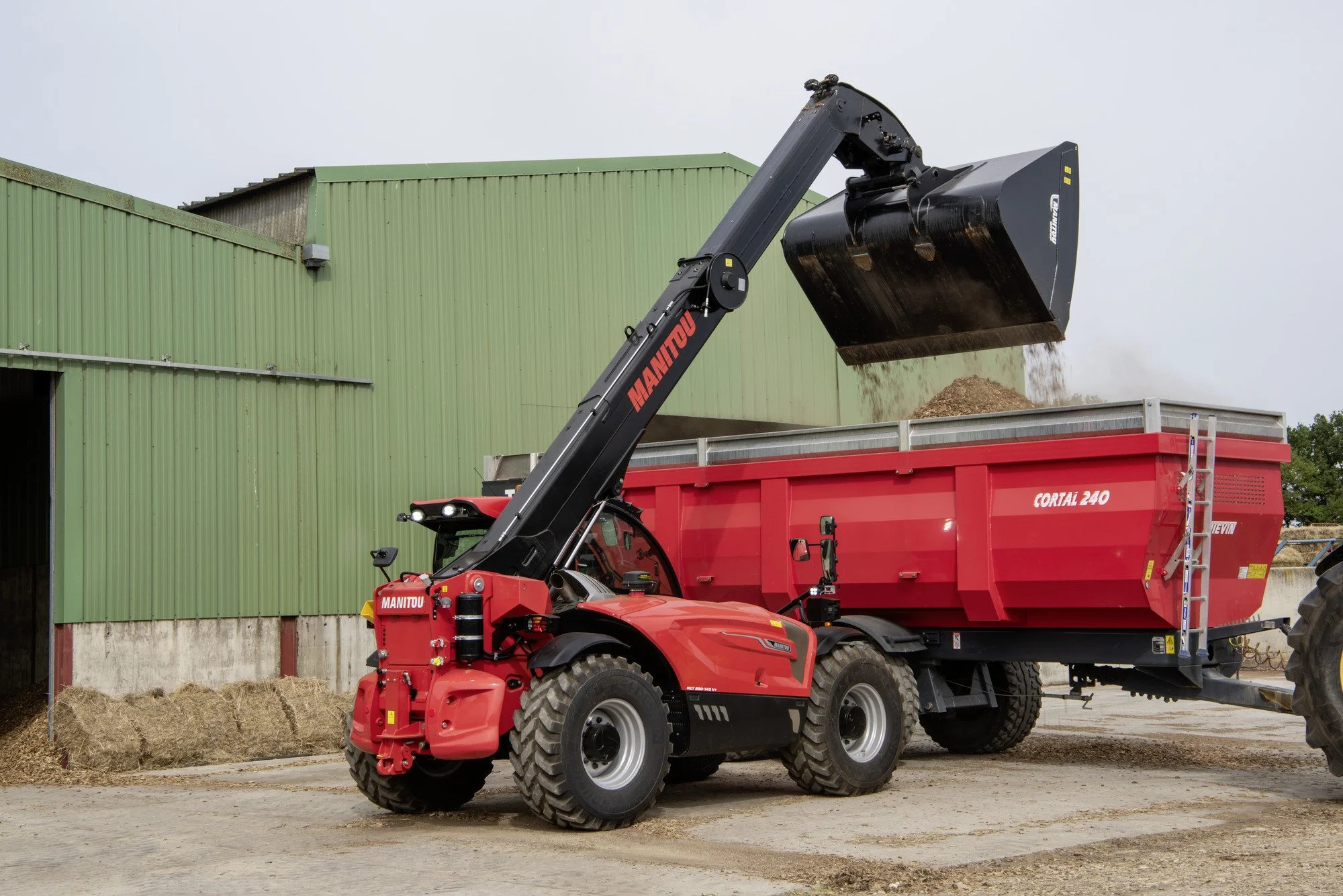 A red Manitou telescopic loader with a black bucket attachment lifting hay into a red trailer, standing outside a green barn on a farm.