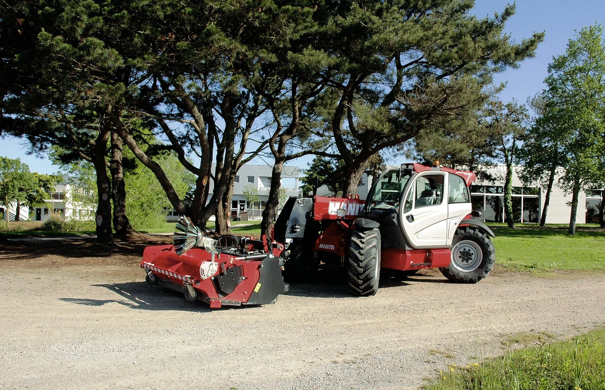 A red and white compact utility tractor with attachments parked on a gravel area under large trees, with modern buildings in the background.