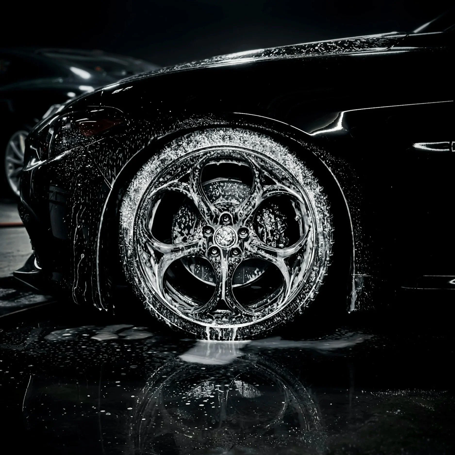 Close-up of a black luxury sports car being washed, with soap suds and water on the wheel and body of the vehicle.