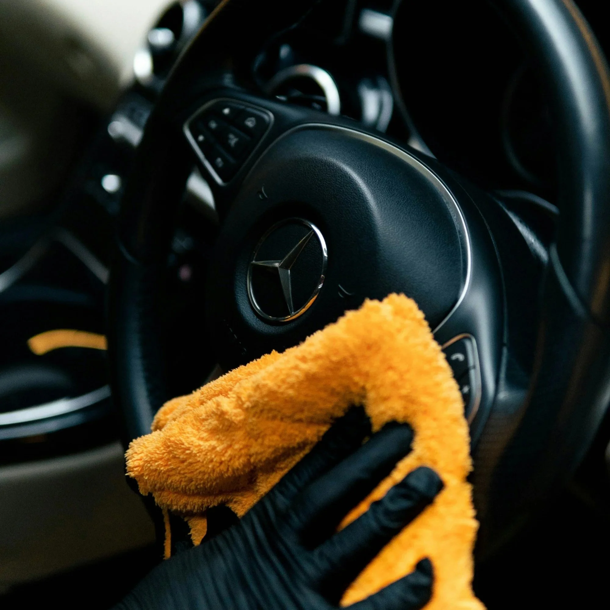 Close-up of a Mercedes-Benz steering wheel with a yellow microfiber cloth in the foreground, and blurred dashboard controls in the background.