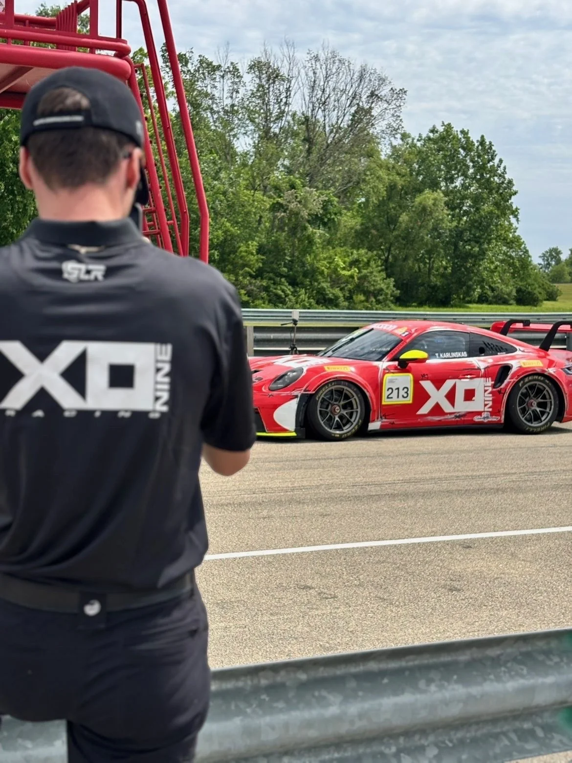 A red racing car with the number 213 on the side and lettering that reads 'XD NINE' is parked on a racetrack. A person wearing a black cap and black clothing with the same logo is standing nearby, looking at the car. The background includes trees and a cloudy sky.