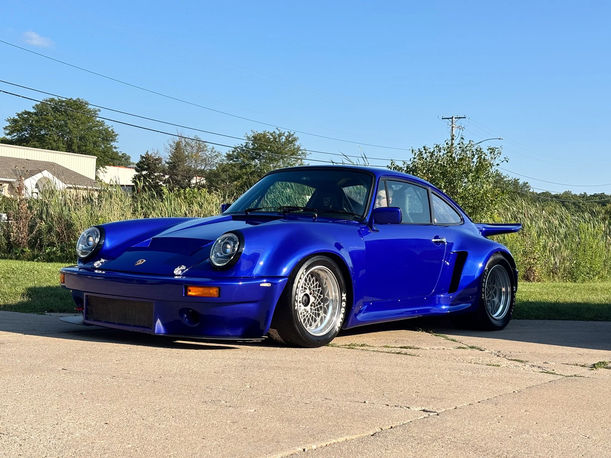 A bright blue Porsche 911 sports car parked on a sidewalk with grass and plants in the background, under a clear blue sky with some clouds and power lines.