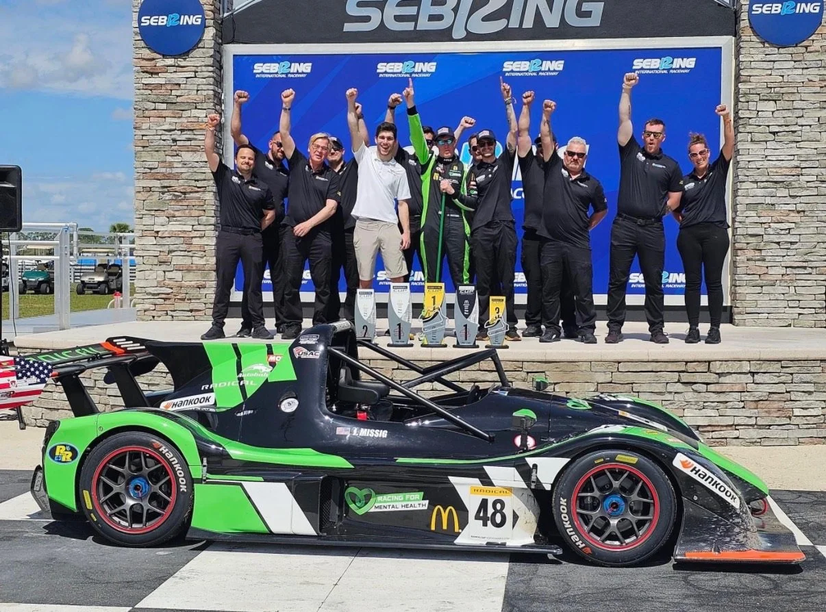 A race car with the number 48 sits on the track in front of a podium. The podium has a group of people celebrating, with some raising their fists. The background features a blue and black backdrop with the SEB Racing logo and branding.