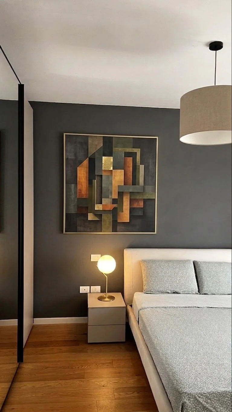 Camera da letto moderna con parete grigia, quadro astratto, lampada da tavolo accesa, letto con coperte chiare, lampadario sopra il letto.