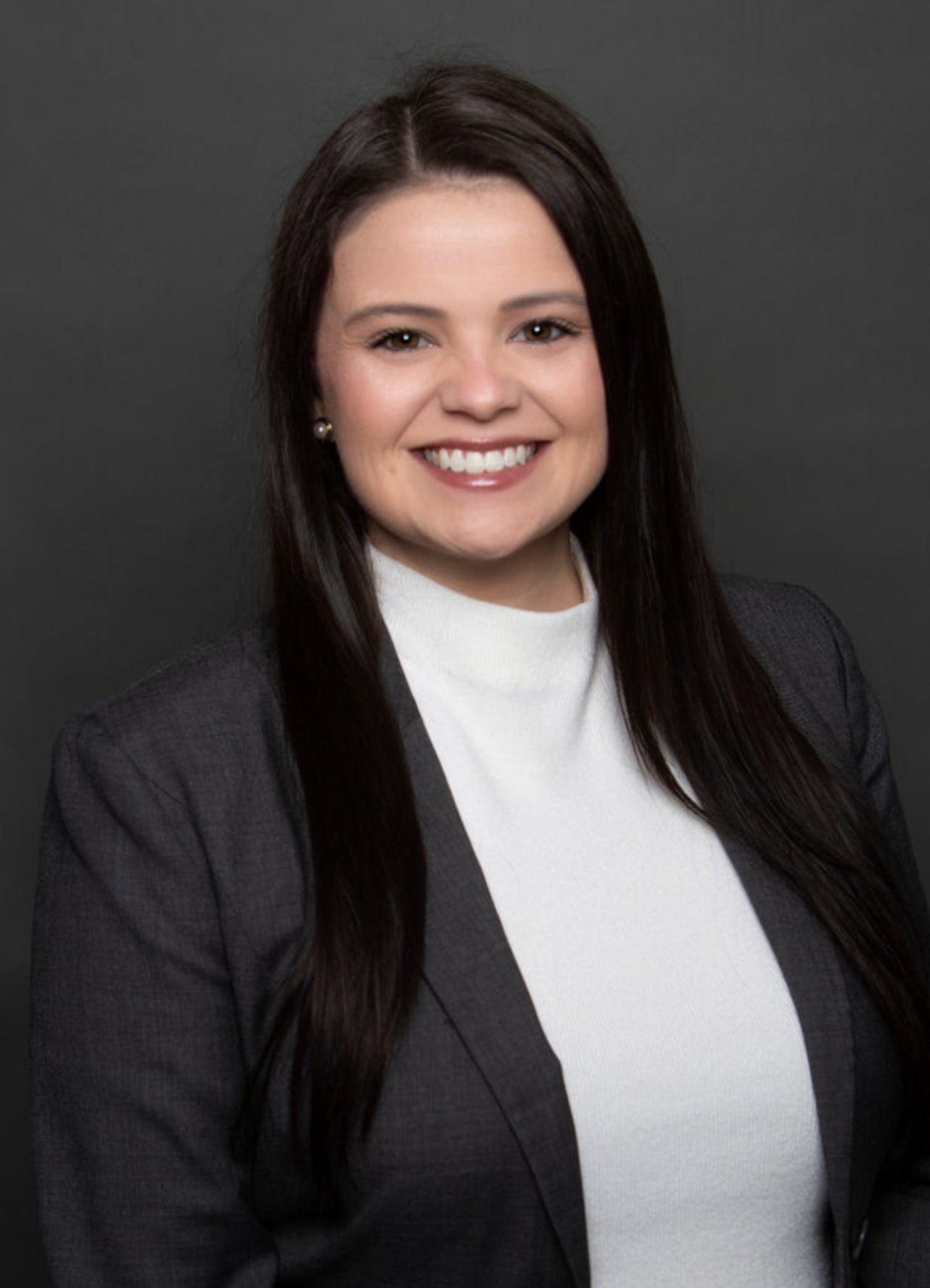 Professional headshot of a woman with long dark brown hair smiling, wearing a grey blazer and white top, against a dark grey background.