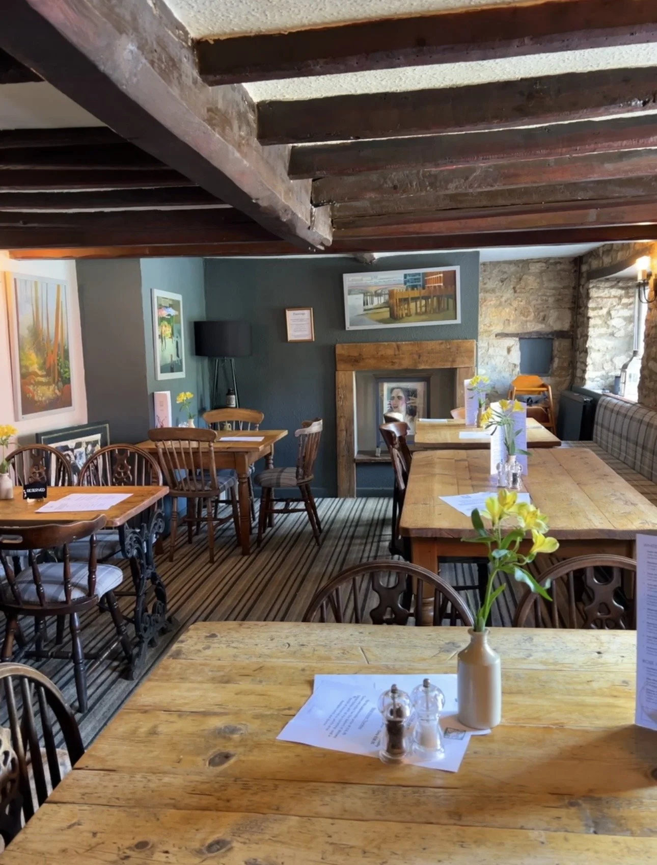 Cozy restaurant interior with wooden tables and chairs, paintings on the walls, a fireplace, flowers on the tables, and natural light from windows.
