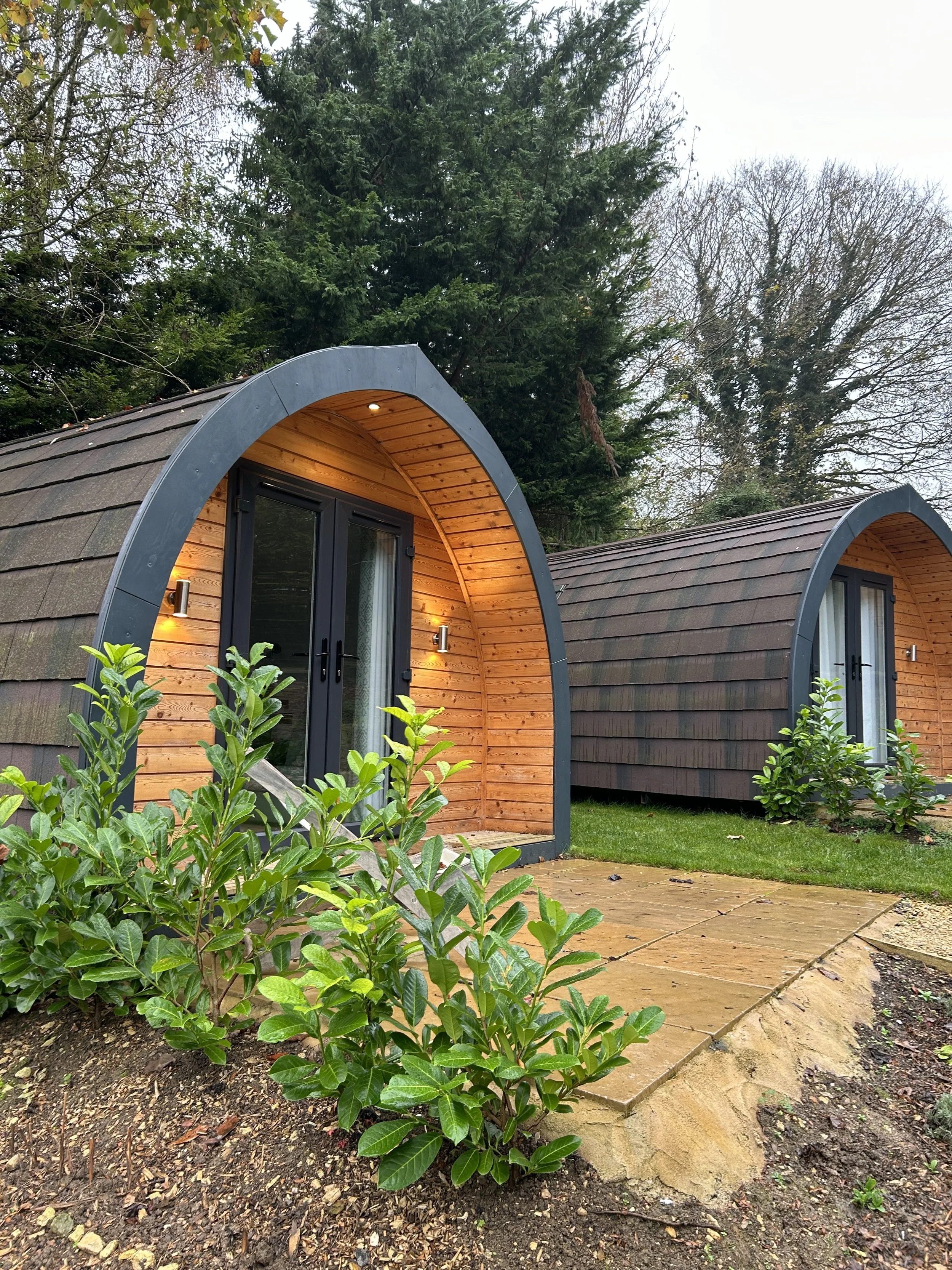 Two small, rounded lodge cabins with wooden exteriors and black doors are situated among trees. One tiny home has a lit entrance with outdoor lights, and there is a stone pathway leading to it. Green shrubs and grass surround the area.