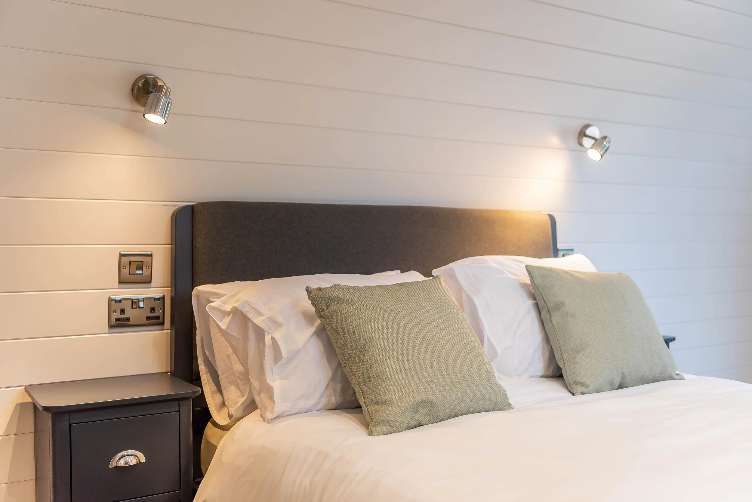 A neatly made bed with white sheets and two beige pillows, a dark gray headboard, and wall-mounted reading lights in a modern bedroom with white wooden paneled walls.