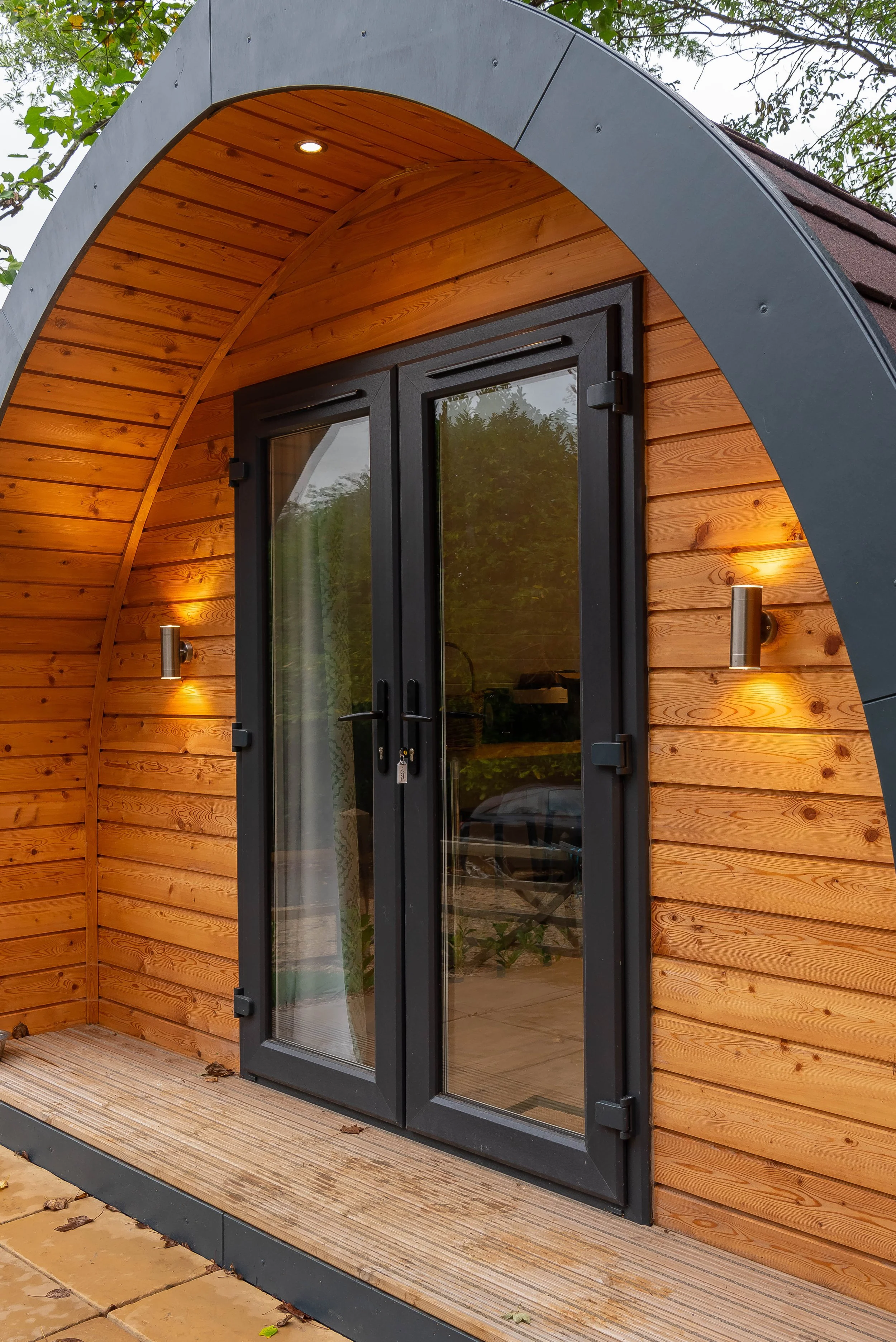 Front of a small wooden house with black double doors, curved roof, outdoor lighting, and wooden deck.