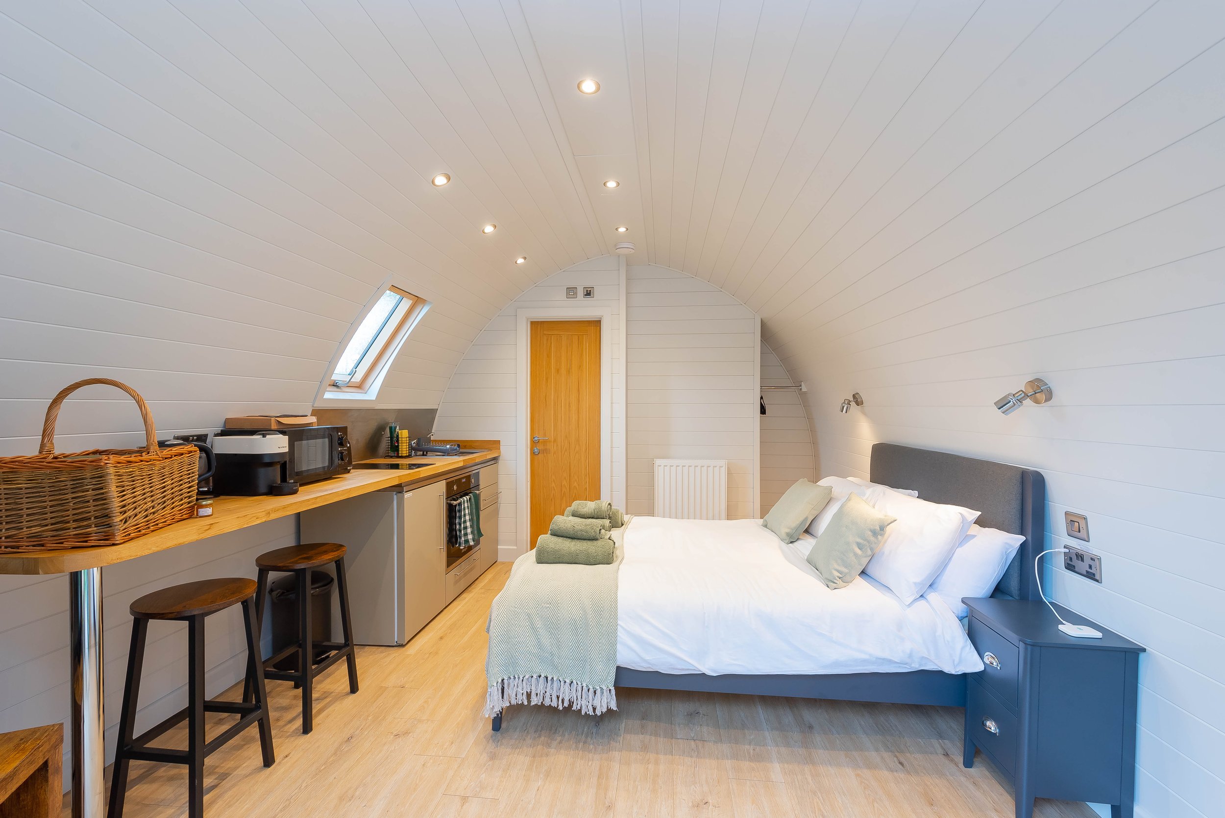 Cozy cabin hotel room with a bed, side table, kitchenette, and skylight window, featuring white walls and wooden accents.