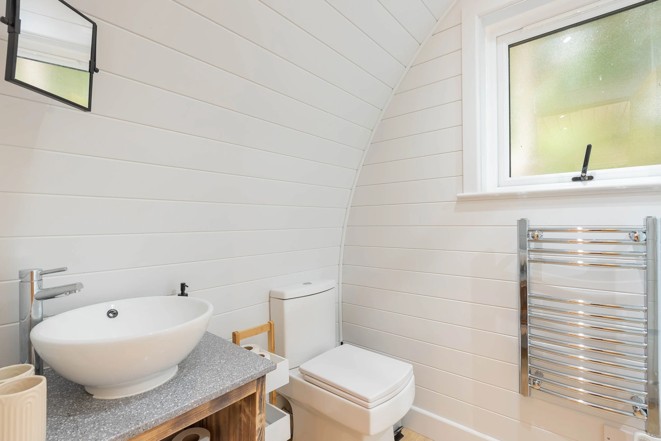 A modern bathroom with white shiplap walls, a small vanity with a vessel sink, a toilet, a window with a view of greenery, and a chrome towel warmer.